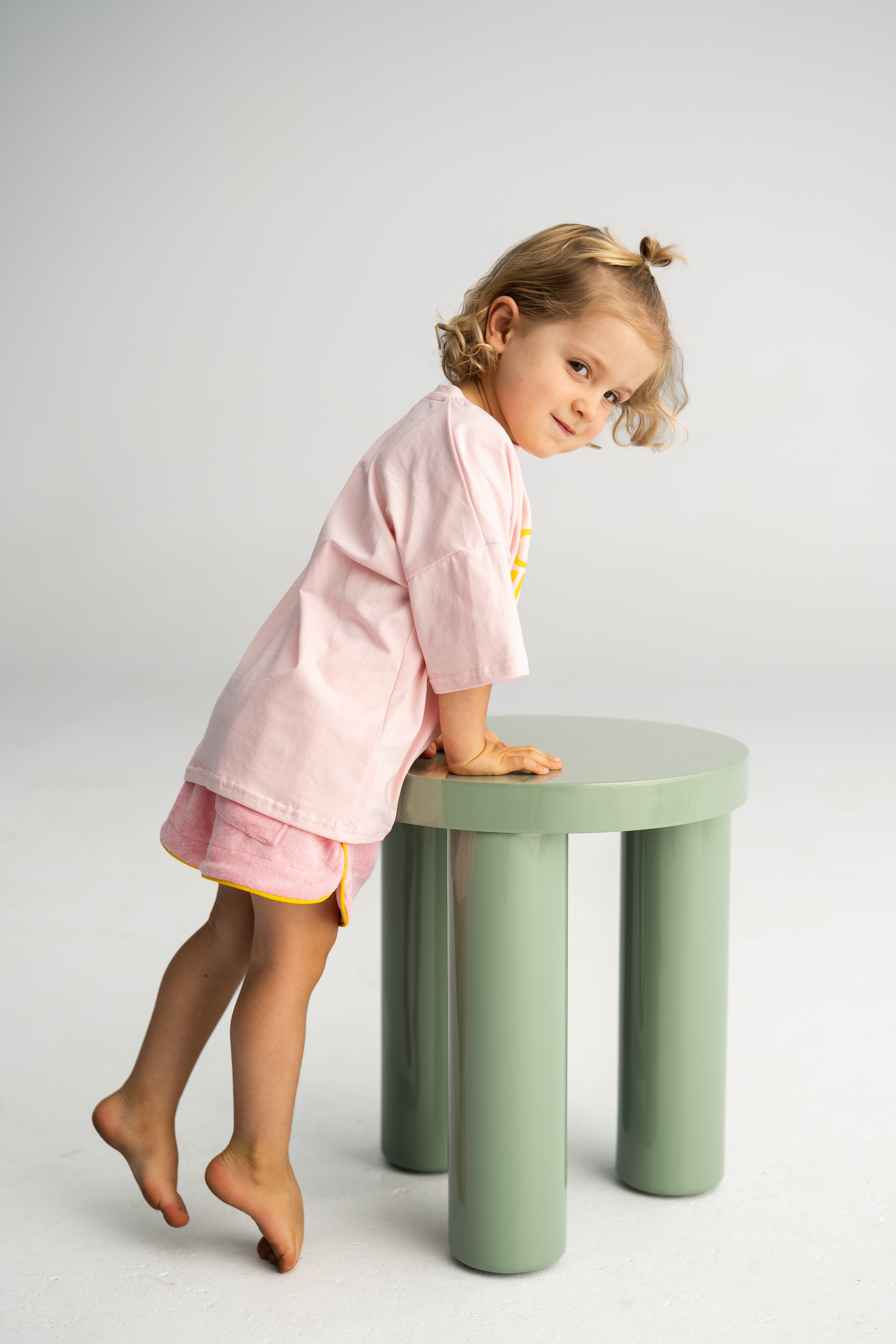 A young child with curly hair, dressed in the SONNY LABEL Terry Shorties Pink loose-fit outfit, leans on a small, round, green stool against a plain light gray background, creating a minimalist setting.