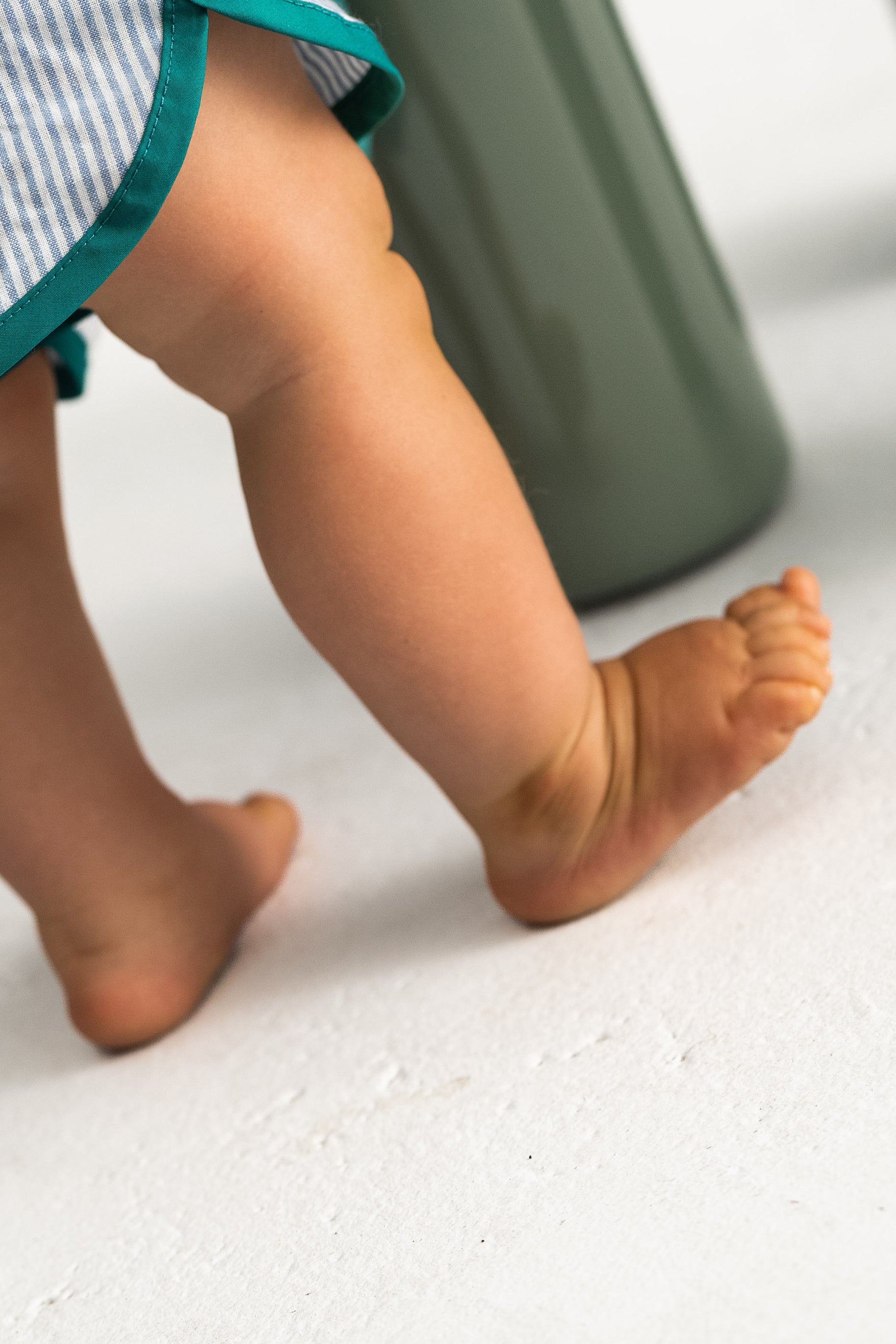 A small child stands barefoot on a light floor, partly wearing SONNY LABEL's Stripe Shorties Blue - Size 6, featuring blue and white stripes with green trim. The child's legs in lightweight shorts are visible as one foot raises slightly, showing chubby toes near a large green object symbolizing summer ease.