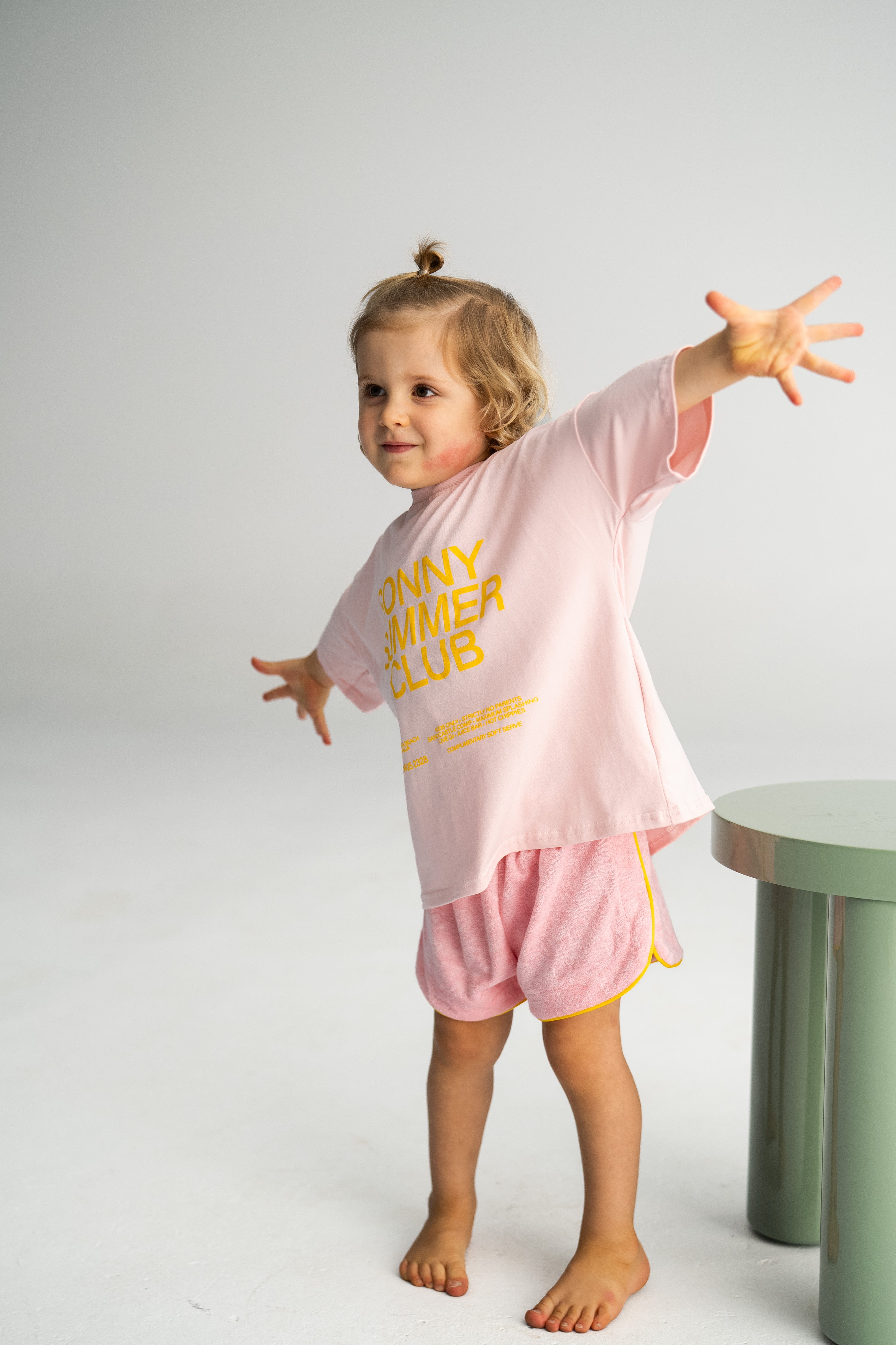 A young child with light brown hair stands on one leg, arms outstretched, wearing a loose-fit pink T-shirt with yellow text paired with Terry Shorties Pink by SONNY LABEL. A green cylindrical object is partially visible on the right, set against a light gray background.