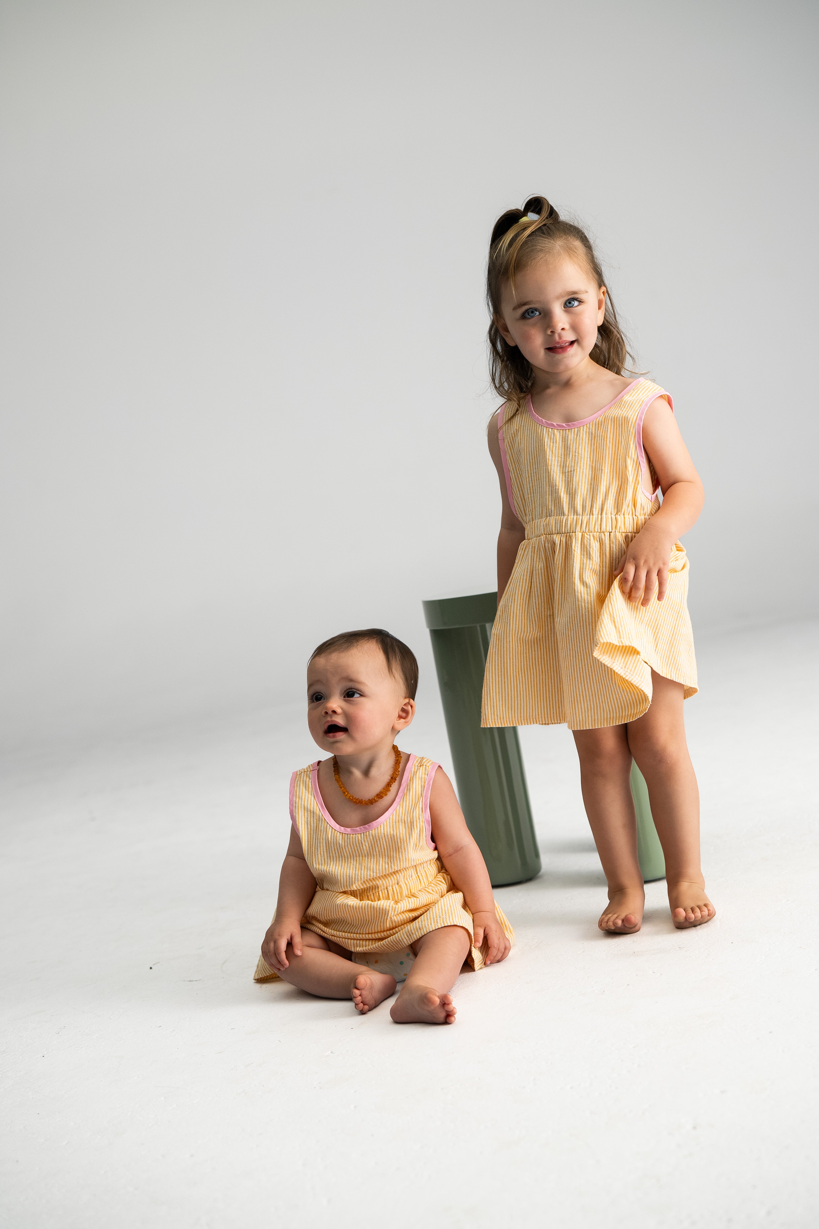 Two young children are wearing matching Stripe Pinafore Lemon dresses by SONNY LABEL. One child sits on the floor while the other stands nearby in a brightly lit, minimalist room adorned with green cylindrical objects.