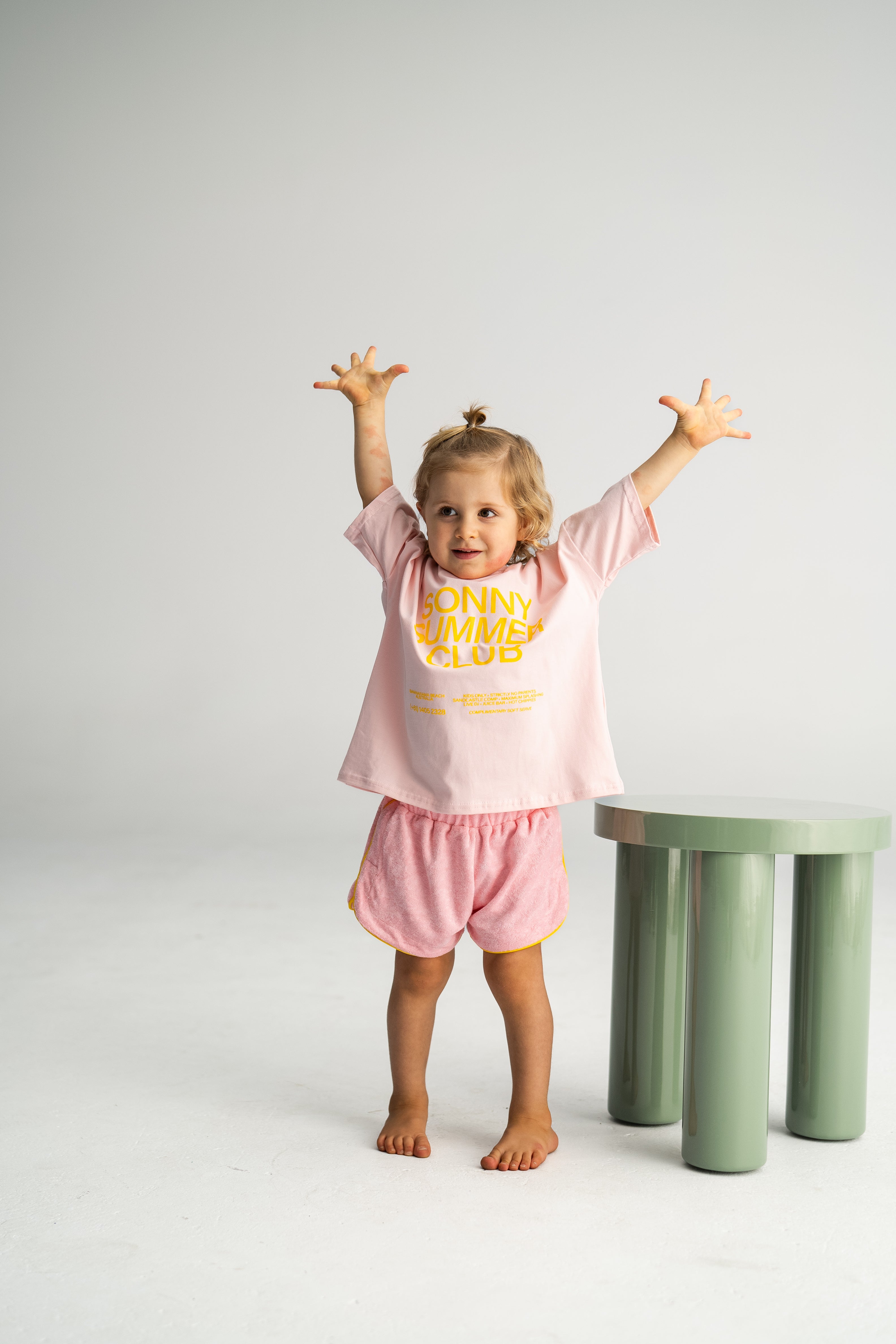 A toddler with light hair stands barefoot on a white floor, arms raised joyfully in a pink T-shirt paired with SONNY LABEL's Terry Shorties Pink. Nearby, a cylindrical green stool complements the plain white background.