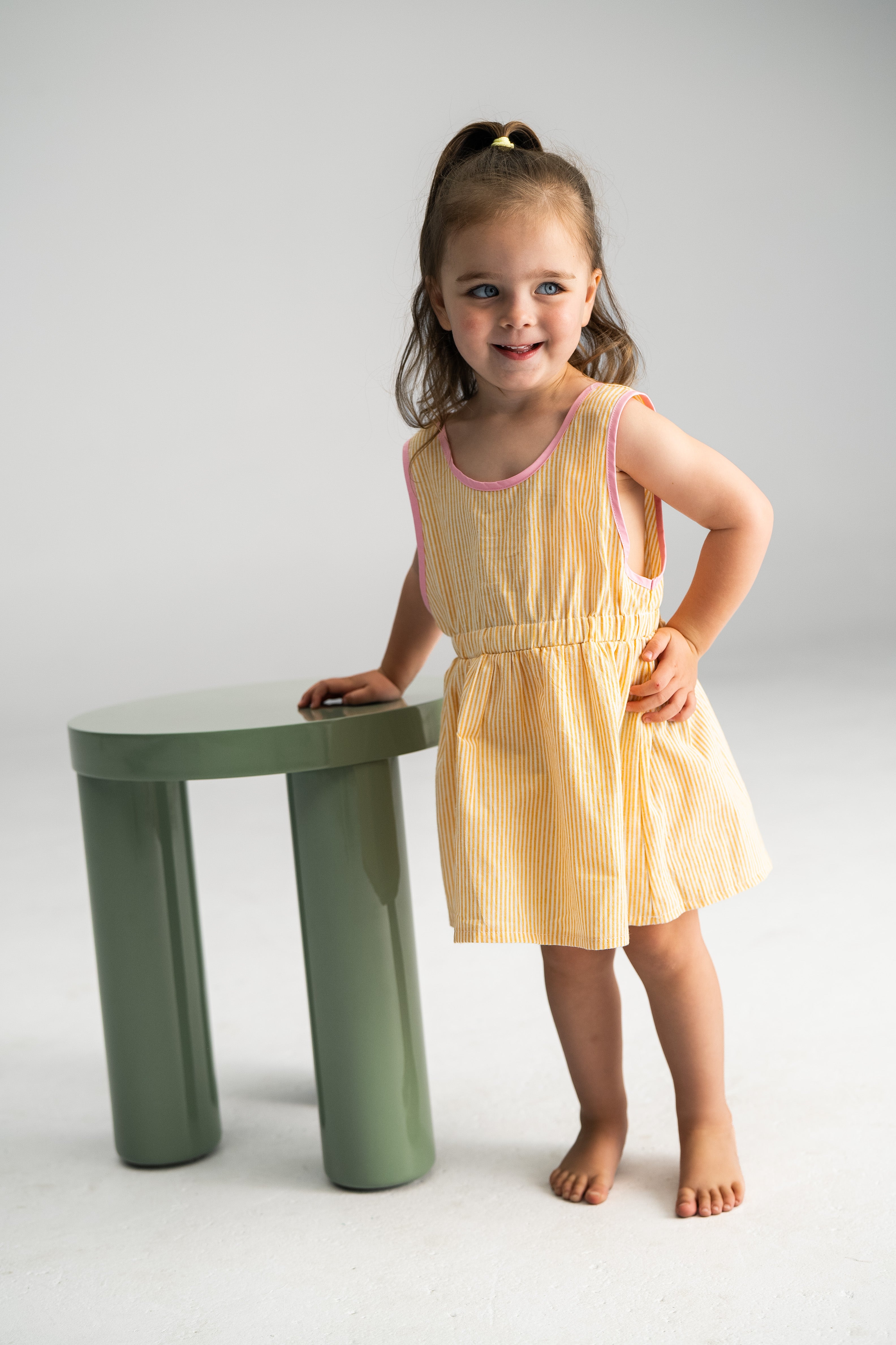 A young girl wearing the Stripe Pinafore Lemon dress from SONNY LABEL stands next to a small green table. Her ponytail bounces as she smiles against a simple, light grey background.