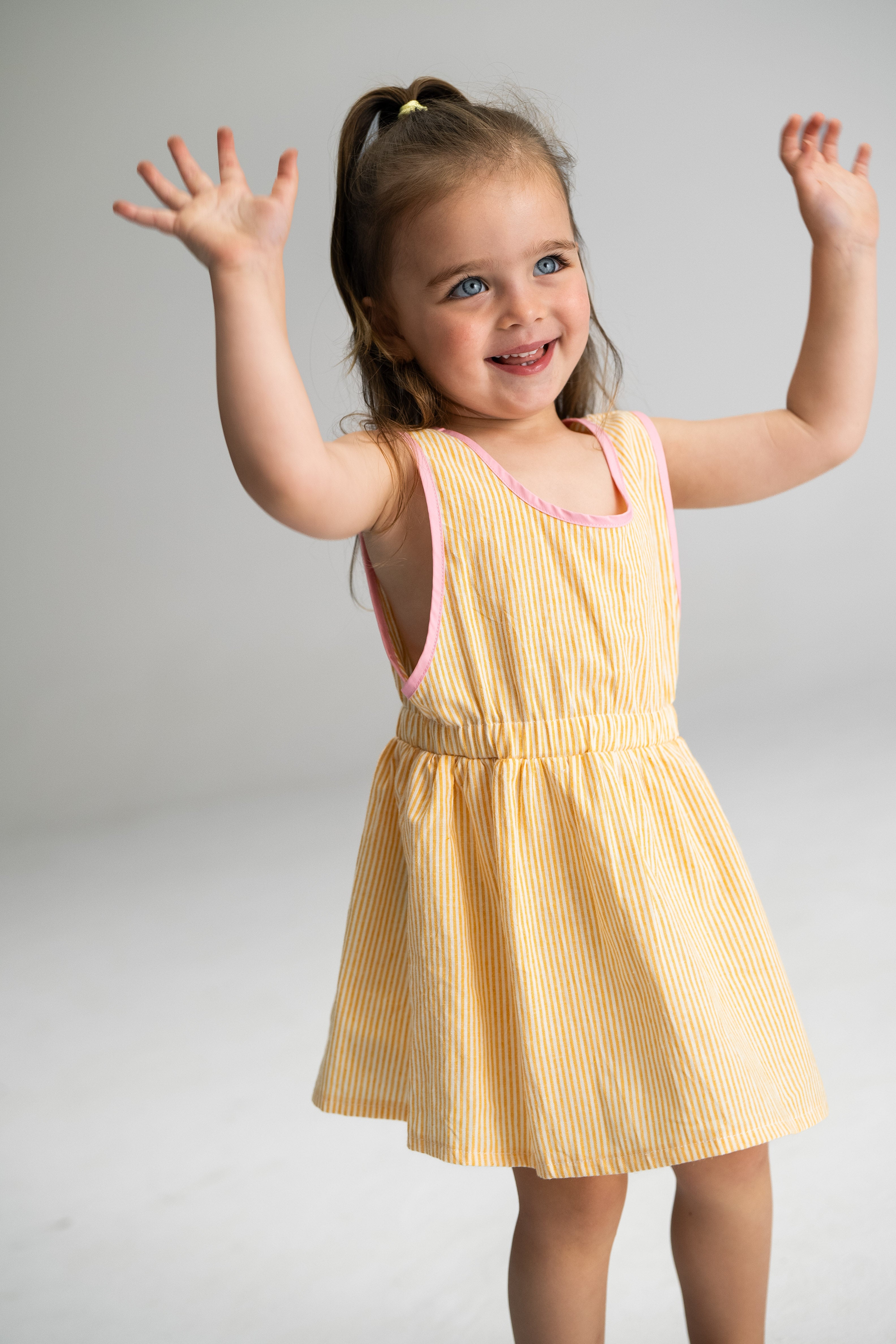 A joyful young girl with brown hair and blue eyes raises her arms in the air, showcasing a delightful Stripe Pinafore Lemon by SONNY LABEL. This cotton dress features yellow and white stripes with pink trim, perfectly complementing her playful spirit as she stands against a plain light gray background.