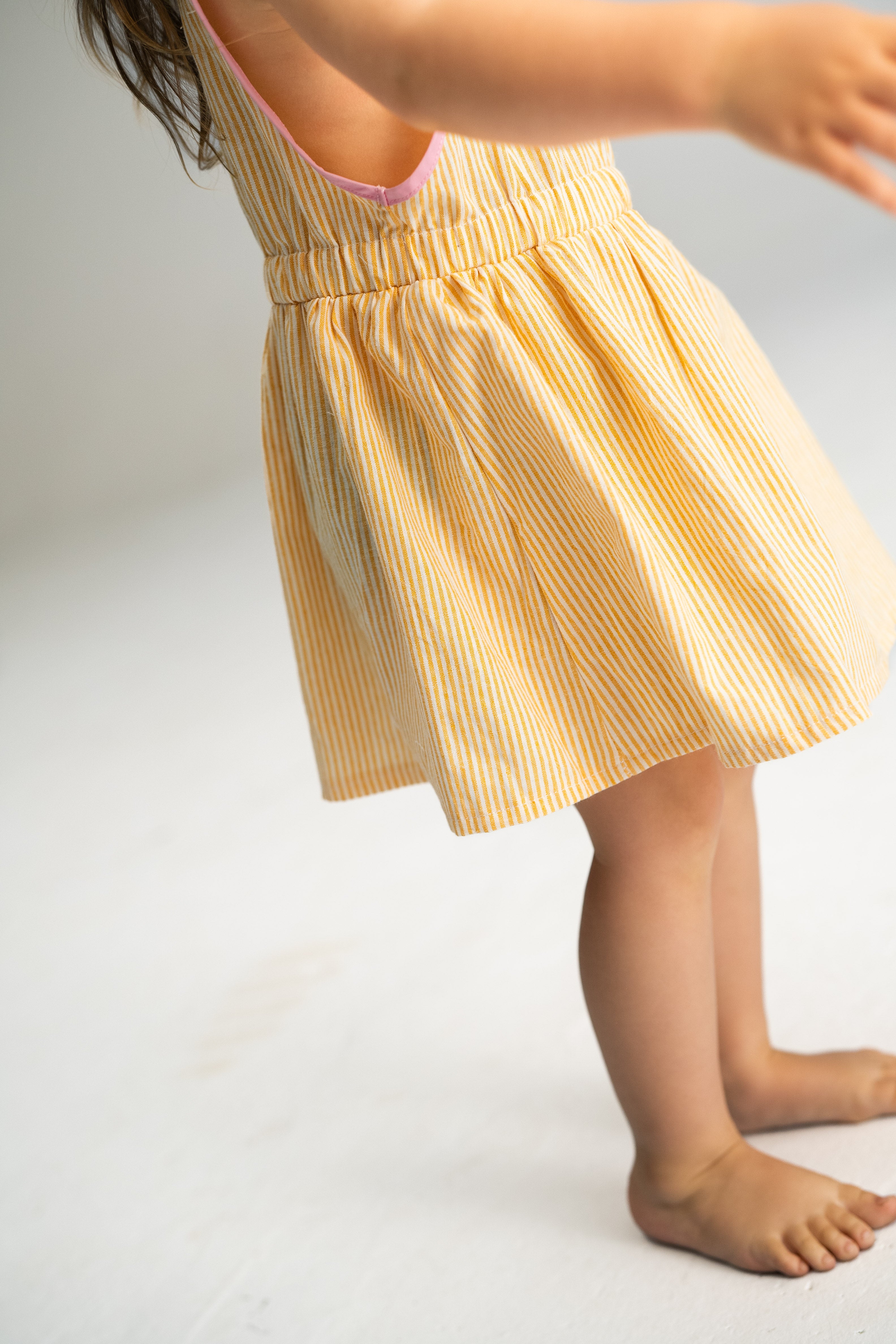 A young child in a sleeveless Stripe Pinafore Lemon dress by SONNY LABEL stands barefoot on a light-colored surface, arms outstretched, with head out of frame. The focus is on the dress and lower body, highlighting one of the charming girls' dresses in playful simplicity.