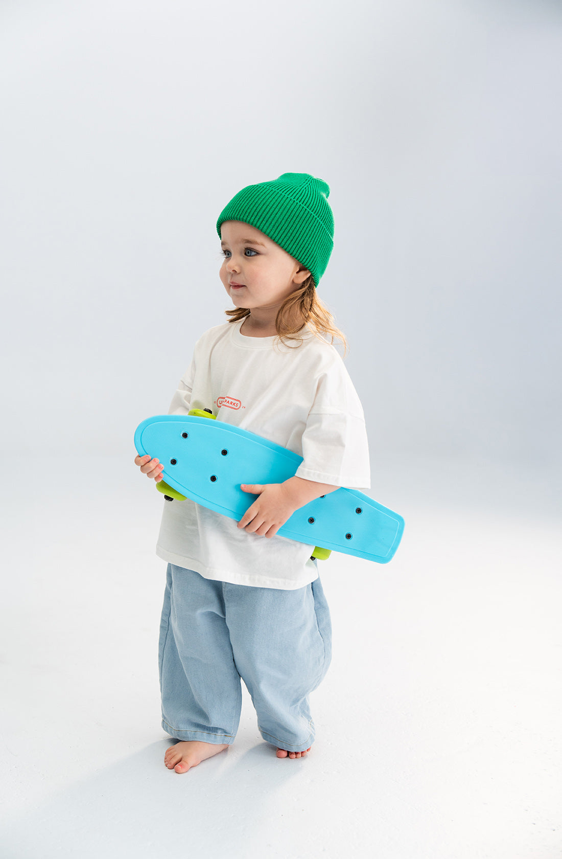 A young child with long hair stands holding a bright blue skateboard. The child is dressed in a loose-fitting white shirt, light blue baggy pants, and a chunky green Cotton Beanie by SONNY LABEL. The white background is simple and uncluttered, giving the scene a clean look.