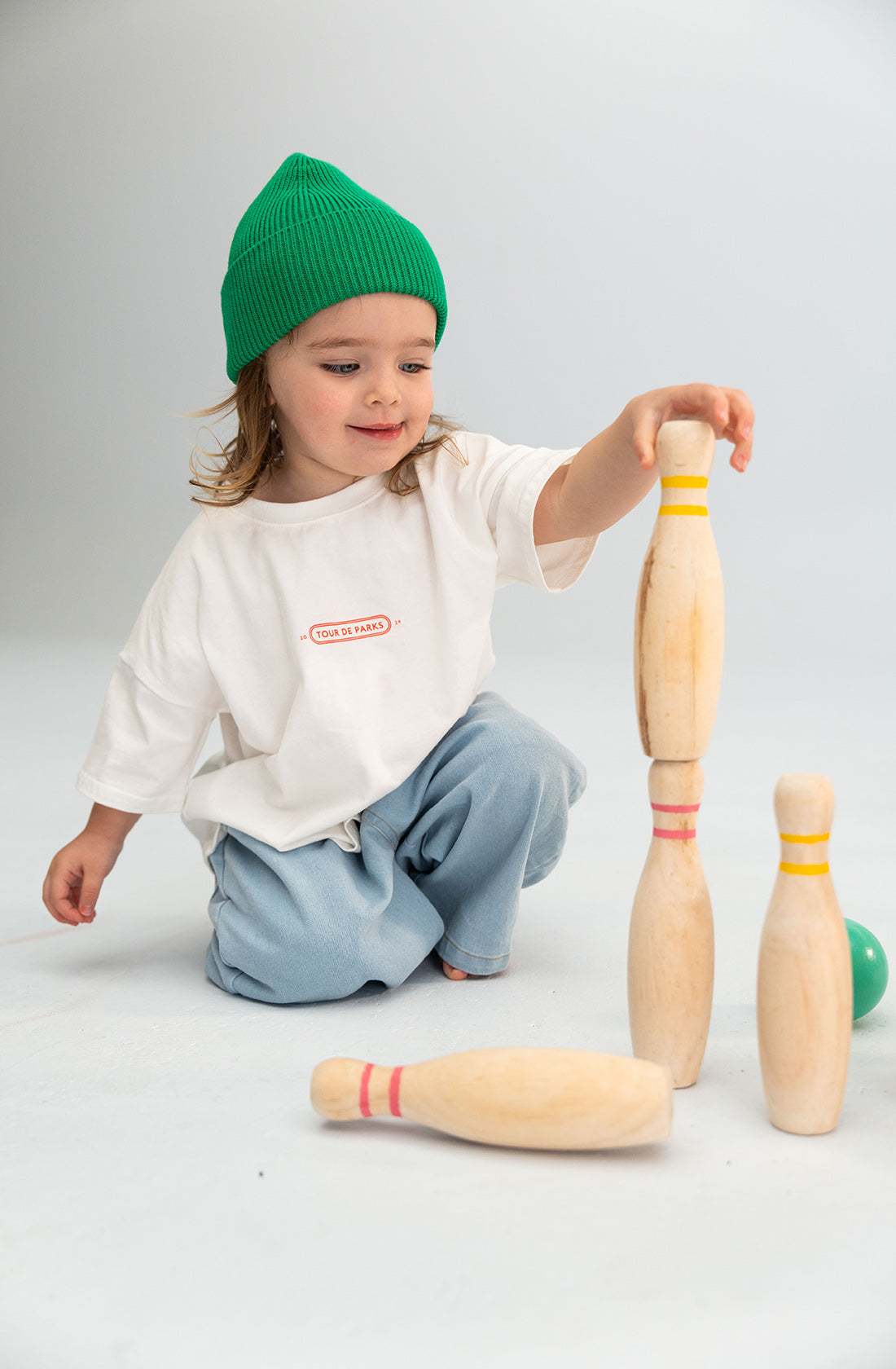 A young child wearing a soft, 100% cotton Cotton Beanie Green from SONNY LABEL, along with a white shirt and light blue pants, is kneeling on the floor and stacking wooden bowling pins. A green ball is placed next to the pins while the background is plain and light-colored.