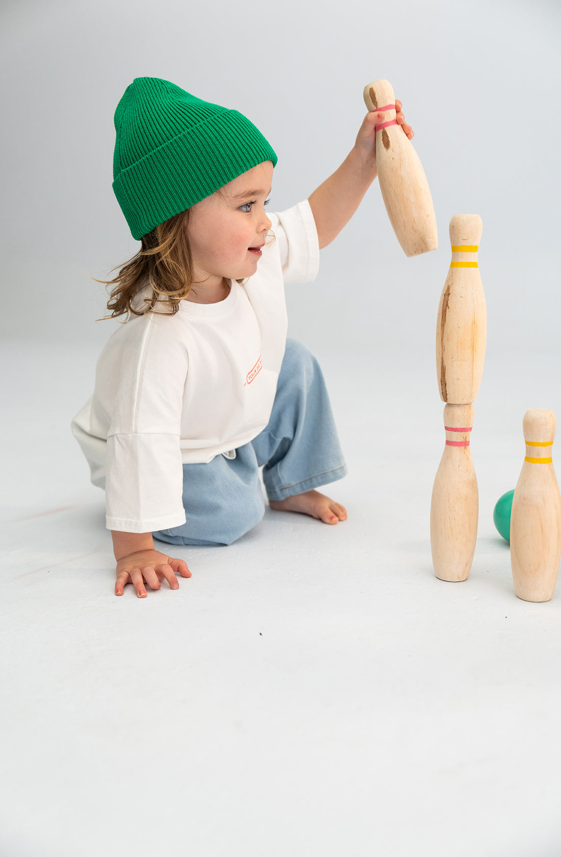 A child wearing a SONNY LABEL Cotton Beanie Green and a white 100% cotton shirt is squatting on the floor, holding a wooden skittle and placing it on top of a stack of other wooden skittles. There is a green ball in the background. The child is focused on balancing the skittles.