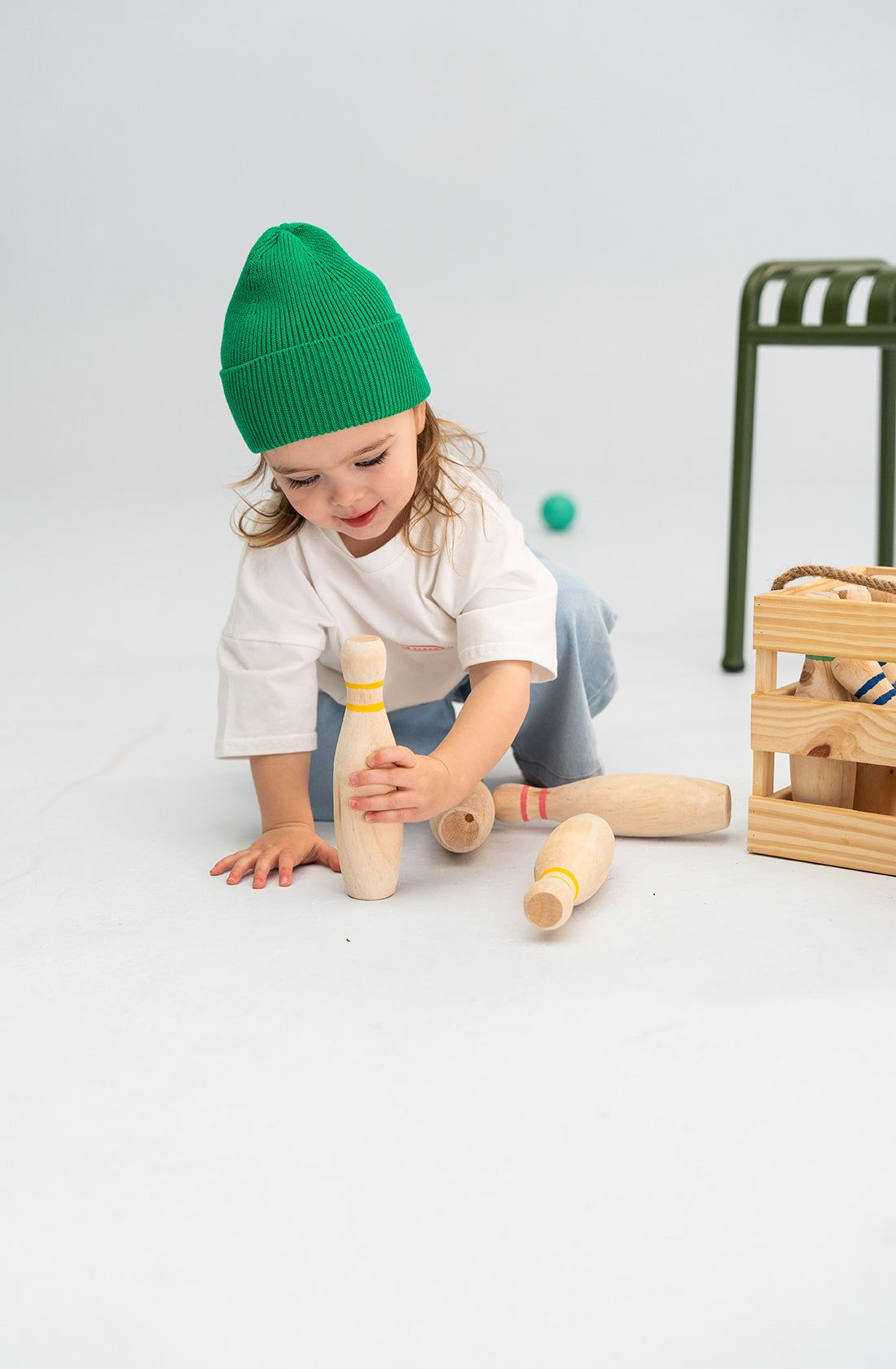 A young child wearing a Cotton Beanie Green from SONNY LABEL and a white t-shirt is playing with wooden bowling pins on the floor. The child is reaching for one of the pins near a wooden crate. A green chair and an additional ball are visible in the background.