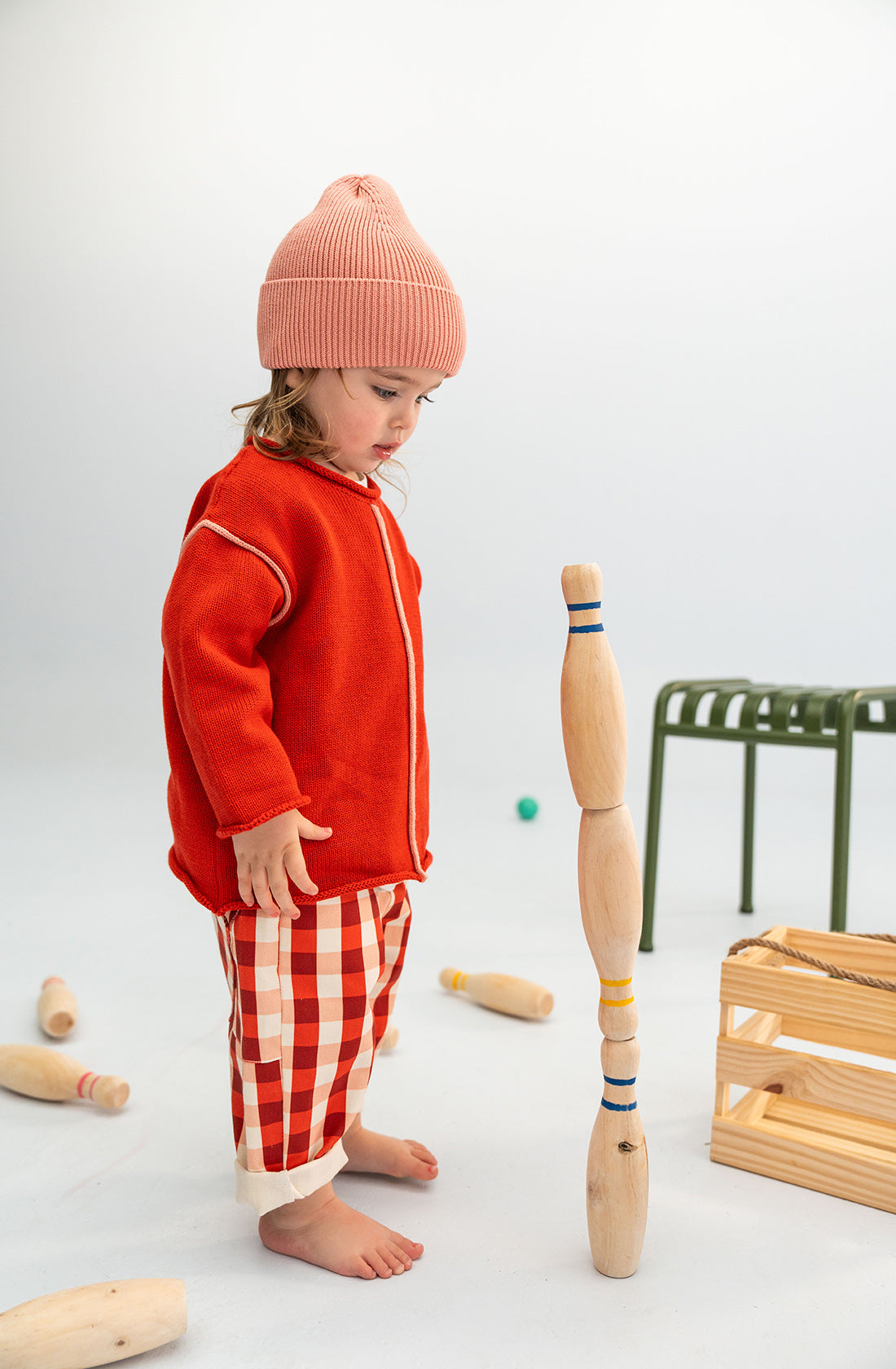 A young child wearing a snuggly soft SONNY LABEL Cotton Beanie Pink, a red jacket, and plaid pants stands barefoot, looking down at a stack of wooden bowling pins. Other pins and a green stool are scattered around on the floor. A wooden crate is also visible to the right.