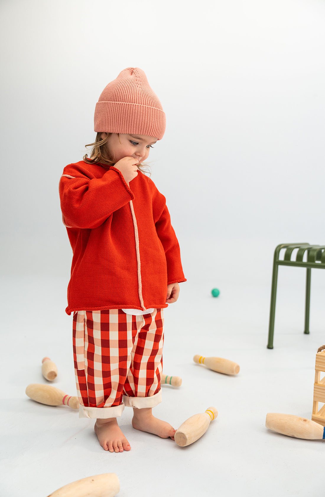 A young child in a comfortably soft Cotton Beanie Pink from SONNY LABEL, a red jacket, and red and white checkered pants stands barefoot on a white floor. The child is looking down and touching their nose while wooden bowling pins lie scattered around, with a green chair in the background.