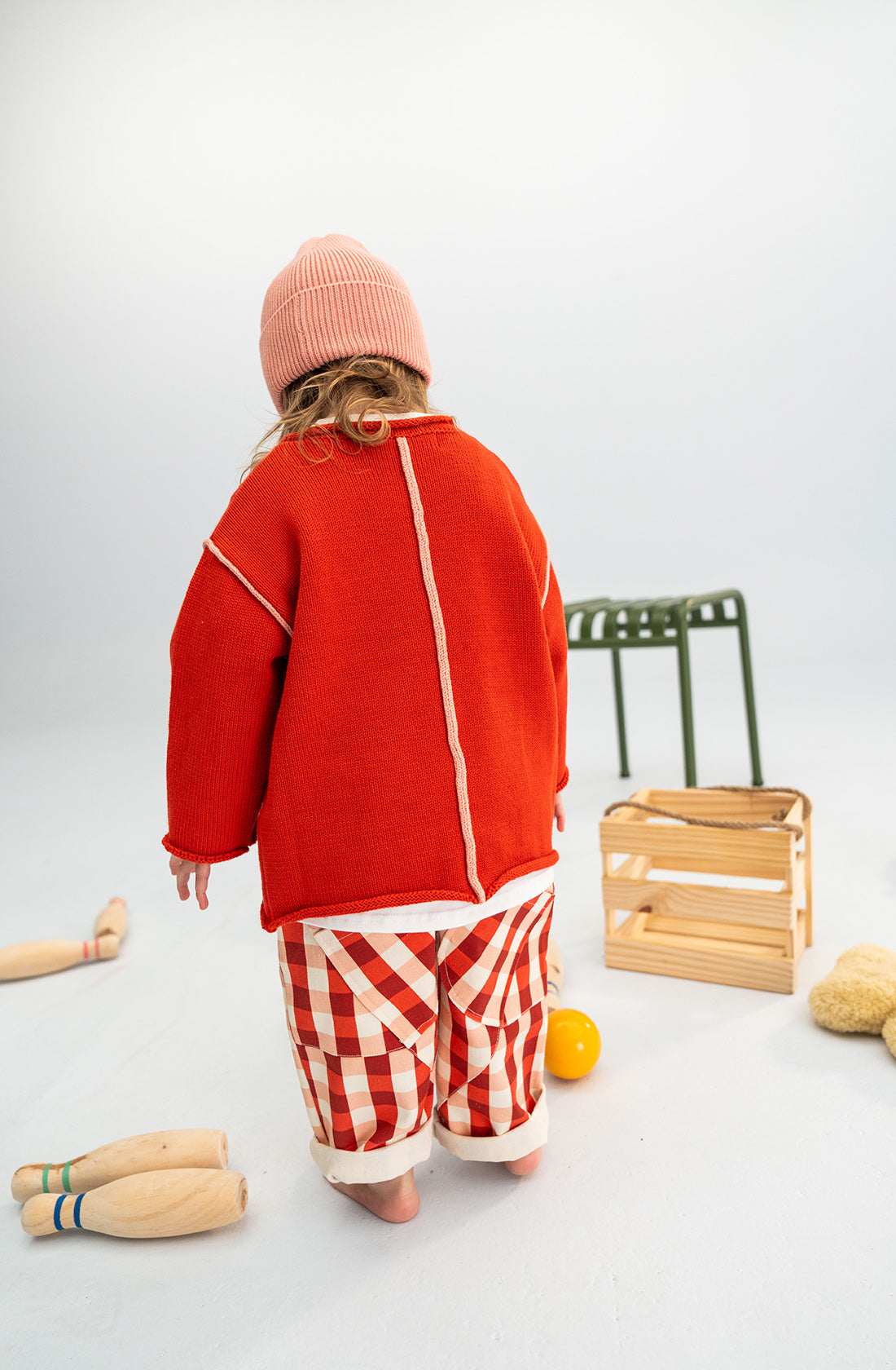 A toddler stands with their back to the camera, wearing a red sweater, red-and-white checkered pants, and a Cotton Beanie Pink by SONNY LABEL. Nearby are wooden bowling pins, a yellow ball, a green chair, and a wooden crate on a white floor with a light gray background.