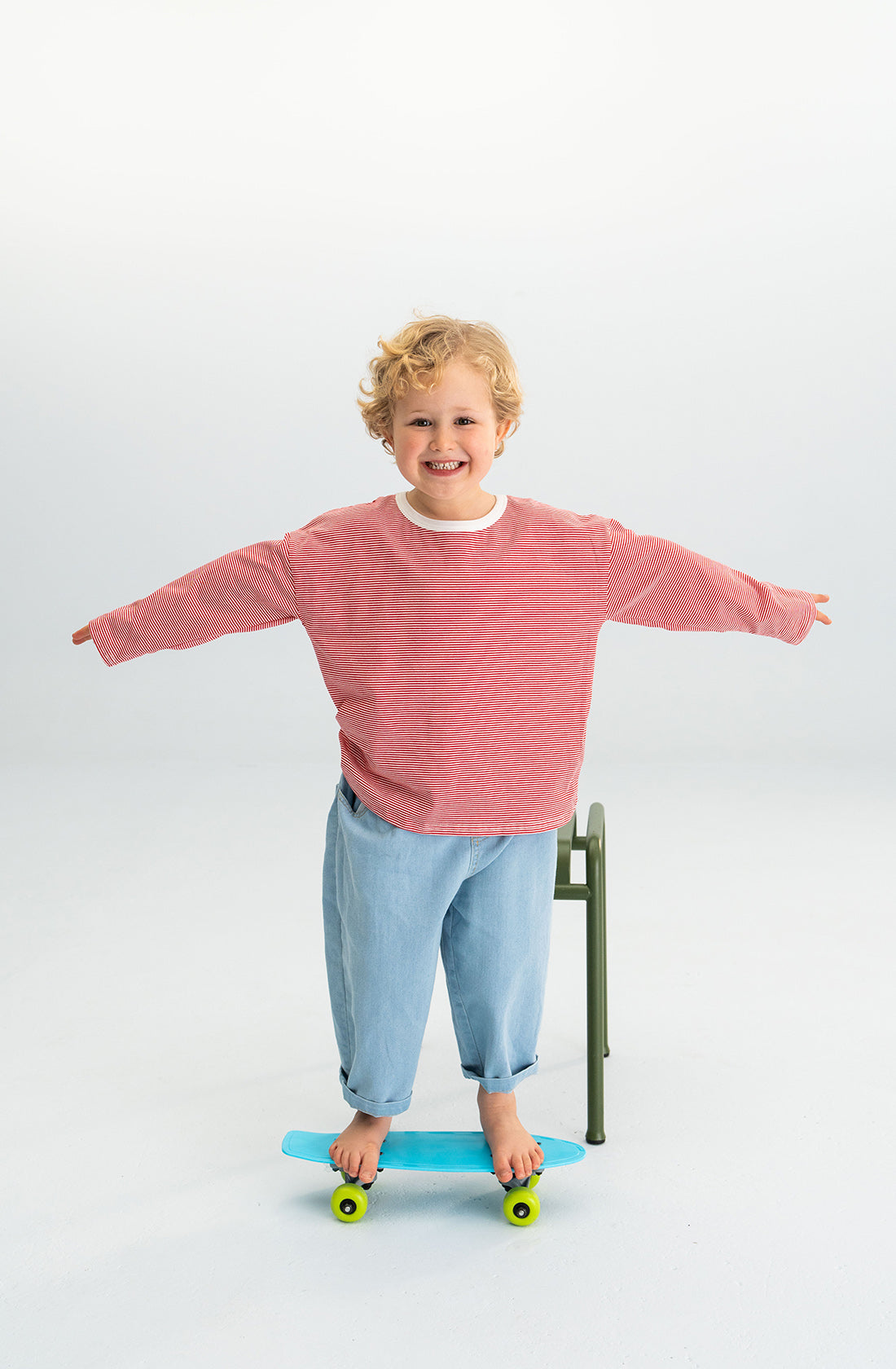 A small child with curly blonde hair stands on a blue skateboard, balancing with arms outstretched. The child is smiling and dressed in a SONNY LABEL Stripe Long Tee Red and light blue jeans, standing on a light-colored floor with a green chair behind them.