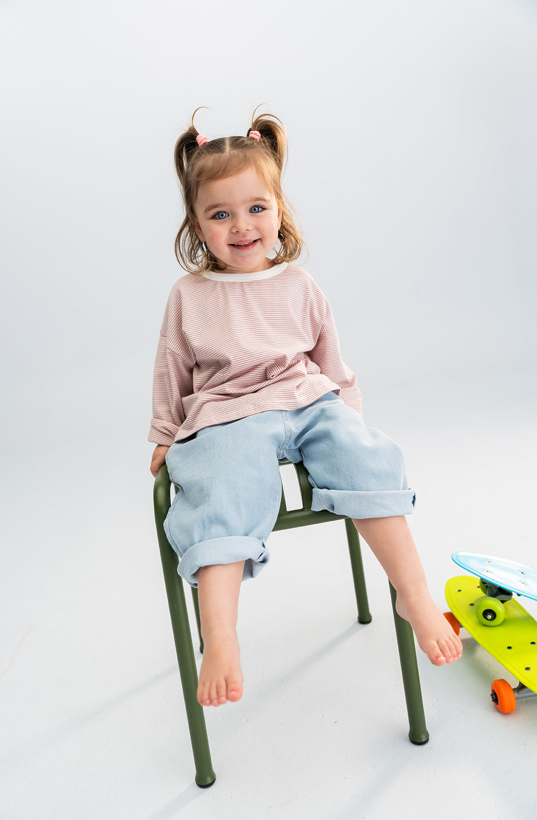 A young child with light brown hair in pigtails sits on a green chair, smiling. The child is wearing a light pink long-sleeve shirt and rolled-up wide leg SONNY LABEL Light Denim Jeans featuring an elastic waistband. A colorful skateboard is placed on the floor to the right. The background is white.