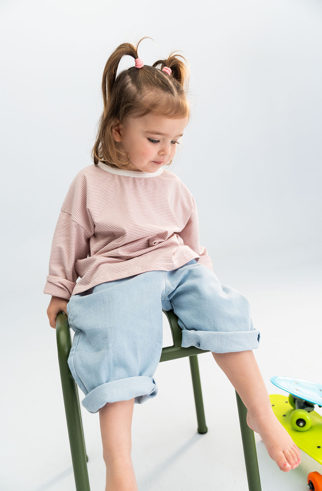 A young girl with two ponytails, wearing a long-sleeved pink shirt and light denim jeans from SONNY LABEL featuring an elastic waistband, sits on a green chair. She gazes downwards with a thoughtful expression, one foot resting on the chair while the other touches the ground.