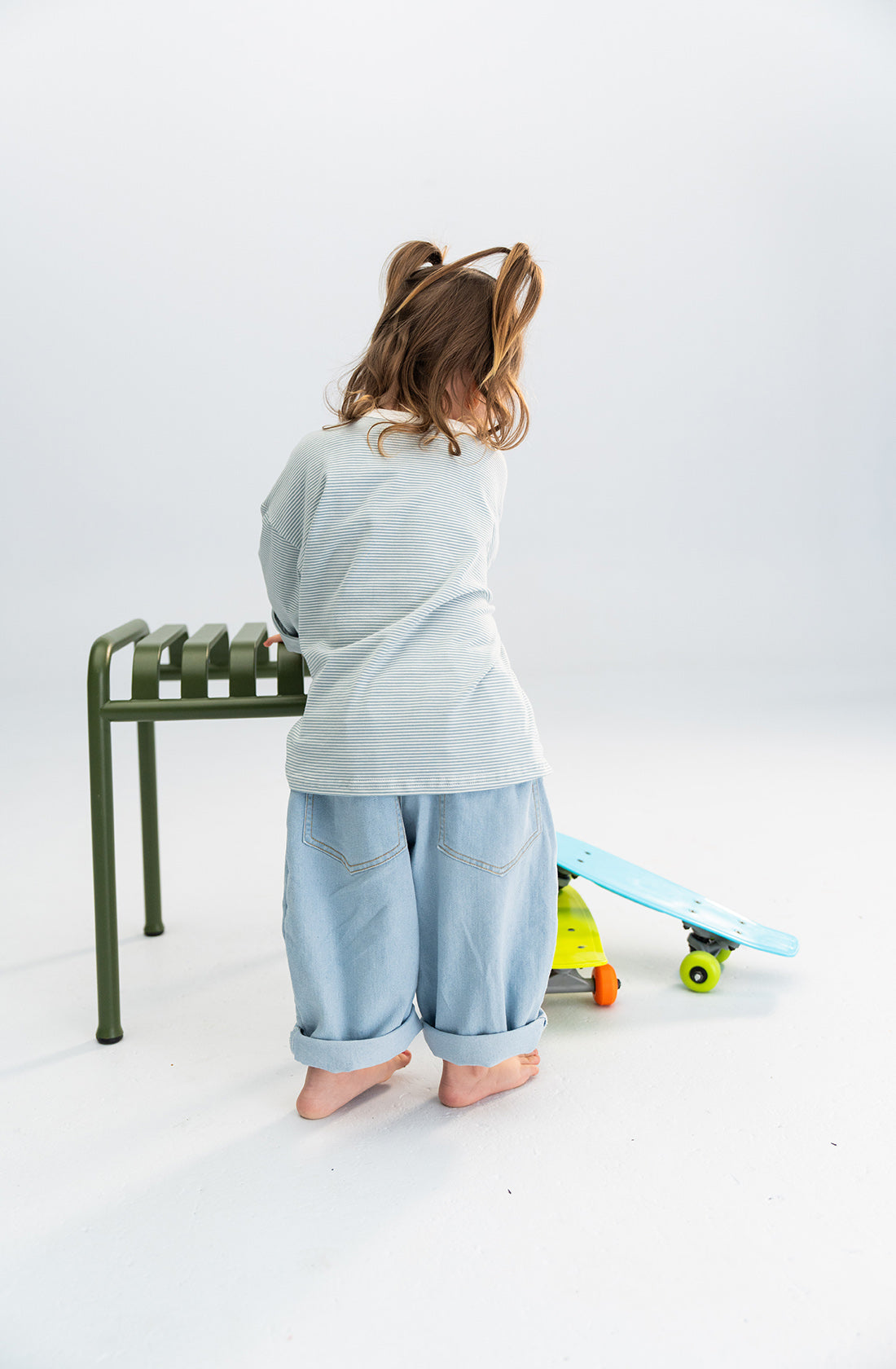 A young child with brown hair in pigtails is standing barefoot, facing away from the camera. They are wearing a SONNY LABEL Stripe Long Tee Blue and loose blue jeans. The child is near a green metal stool and a colorful toy skateboard on the floor, embodying everyday essentials.