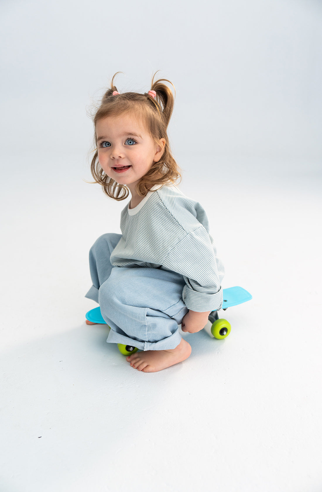 A young child with light brown hair tied in pigtails, wearing a striped long-sleeve shirt and SONNY LABEL's Light Denim Jeans with an elastic waistband, is sitting on a small colorful skateboard. The child is barefoot and has a joyful expression, looking directly at the camera.