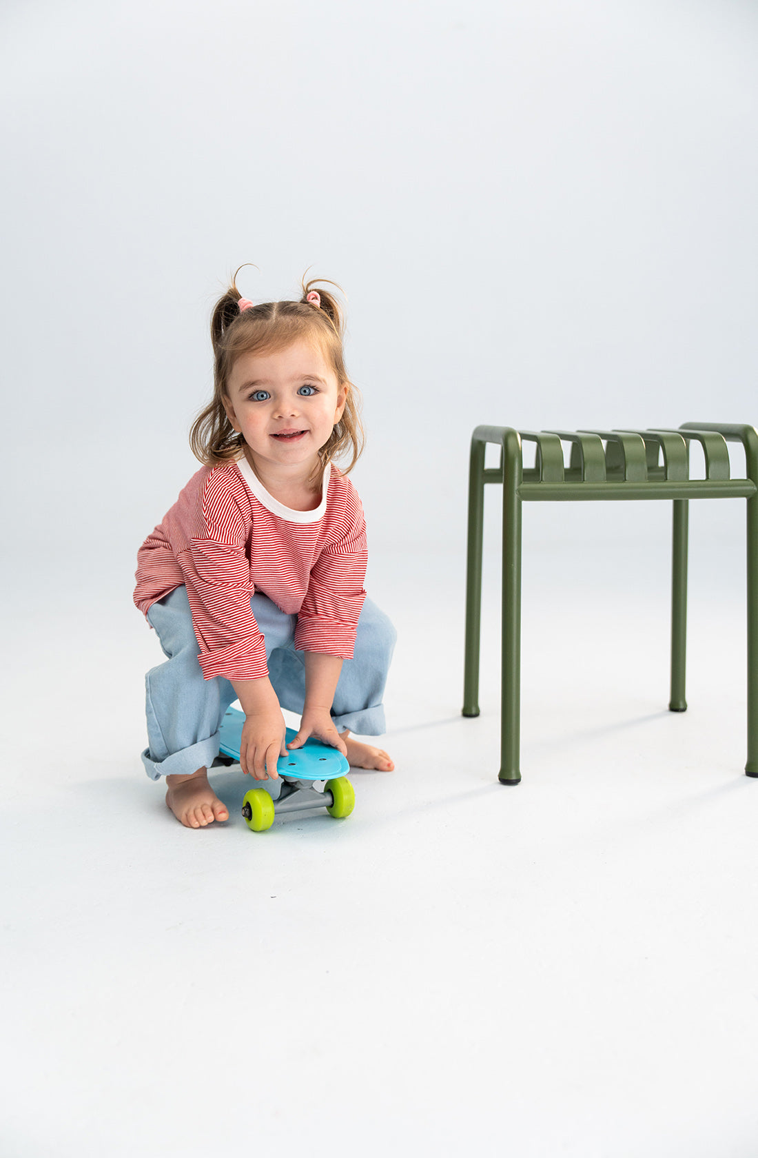 A young child with two small ponytails, wearing a red and white striped long-sleeve shirt and SONNY LABEL Light Denim Jeans featuring an elastic waistband, is smiling while crouching on a small blue skateboard. A green chair is nearby on a white floor with a plain white background.