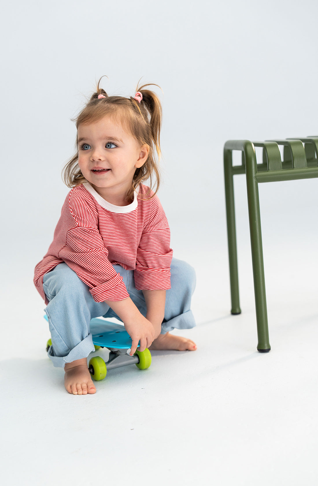 A toddler with pigtails and a Stripe Long Tee Red by SONNY LABEL, an essential piece in any playful wardrobe, sits on a small skateboard. They are barefoot and wearing light blue pants, smiling and looking to the left. Next to them is a green metal chair on a white background.