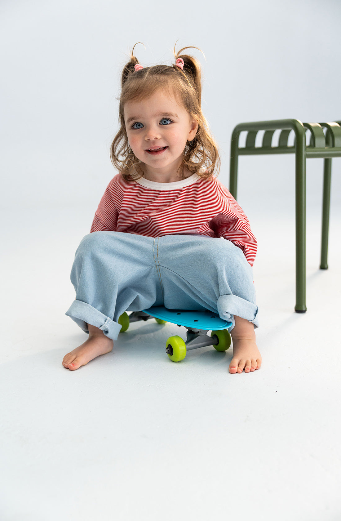 A young child with blond hair in pigtails sits on a small blue skateboard with neon green wheels. Wearing the Stripe Long Tee Red by SONNY LABEL and light blue jeans, the child smiles while looking ahead. An essential piece of home decor, a green metal stool, is seen to the right in the background.