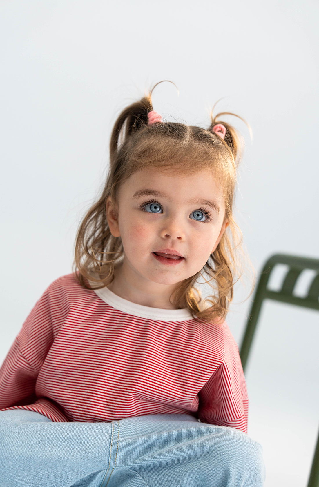 A young child with light brown hair in pigtails and blue eyes is seated on a chair. They are wearing the Stripe Long Tee Red by SONNY LABEL, made of soft fabric, paired with light blue pants. The background is light and uncluttered, drawing attention to the child's expressive face.