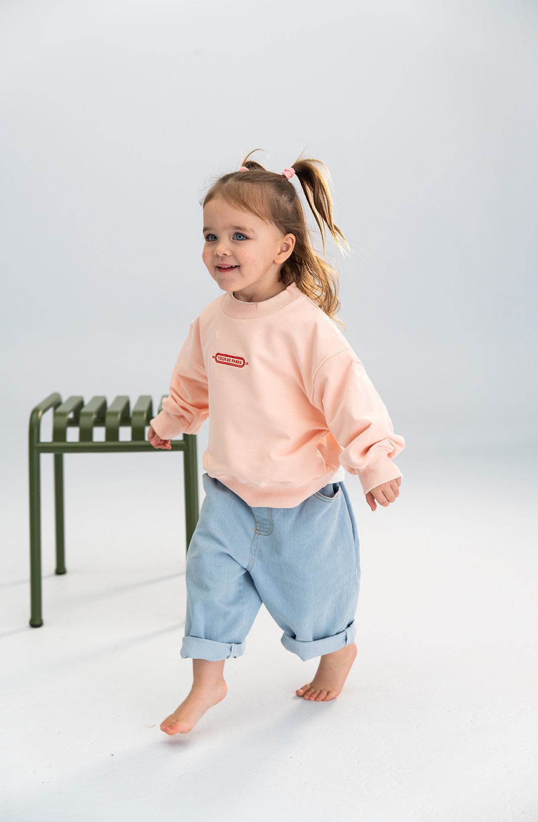 A young child with light brown hair in pigtails is walking barefoot indoors. They are wearing a peach-colored sweatshirt with a small red logo on the chest and SONNY LABEL Light Denim Jeans with an elastic waistband. There is a green metal bench in the background.