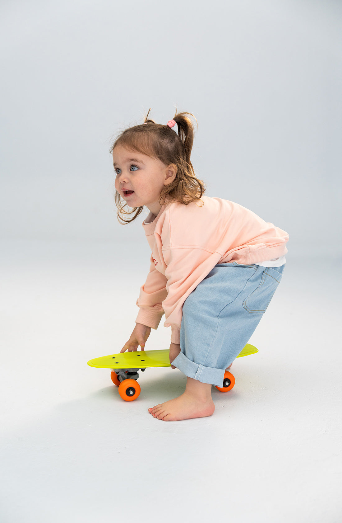 A young child with brown hair in pigtails, dressed in a light pink long-sleeve shirt and SONNY LABEL Light Denim Jeans featuring an elastic waistband, squats while holding a yellow skateboard with orange wheels. The child is barefoot and appears focused, set against a light, plain background.