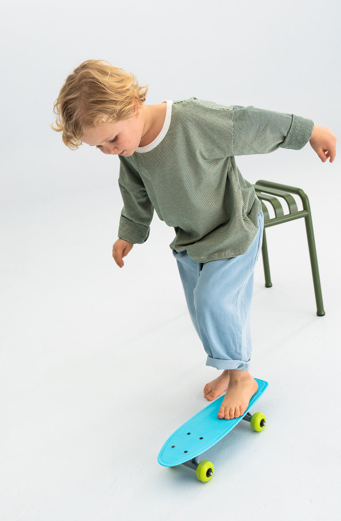 A young child with blonde hair balances on a blue skateboard with yellow wheels. The child is barefoot, wearing the Stripe Long Tee Green from SONNY LABEL, which features bright stripes on the sleeves, paired with light blue pants. Using a green metal stool for support, this scene captures an everyday essential moment of playful exploration.