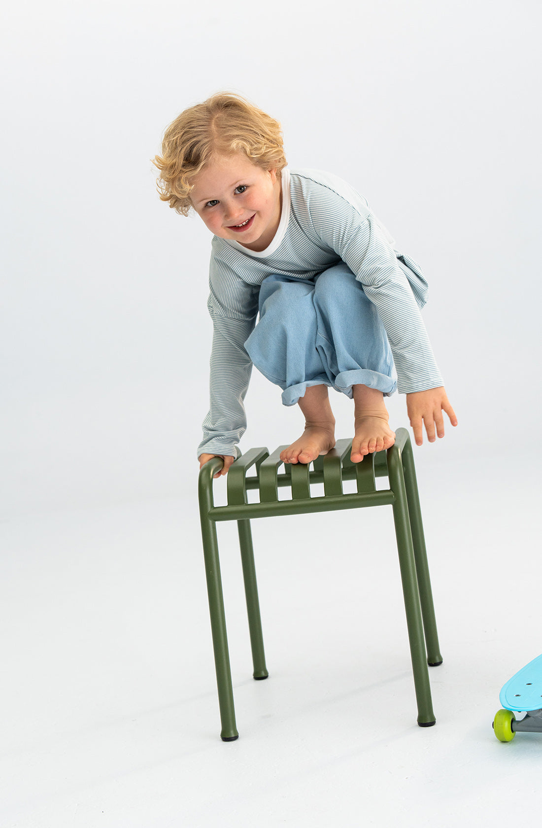 A child with curly blond hair is crouching on top of a small green stool, smiling and balancing. The child wears the Stripe Long Tee Blue by SONNY LABEL and light blue pants. A blue skateboard is partially visible on the right side of the image.