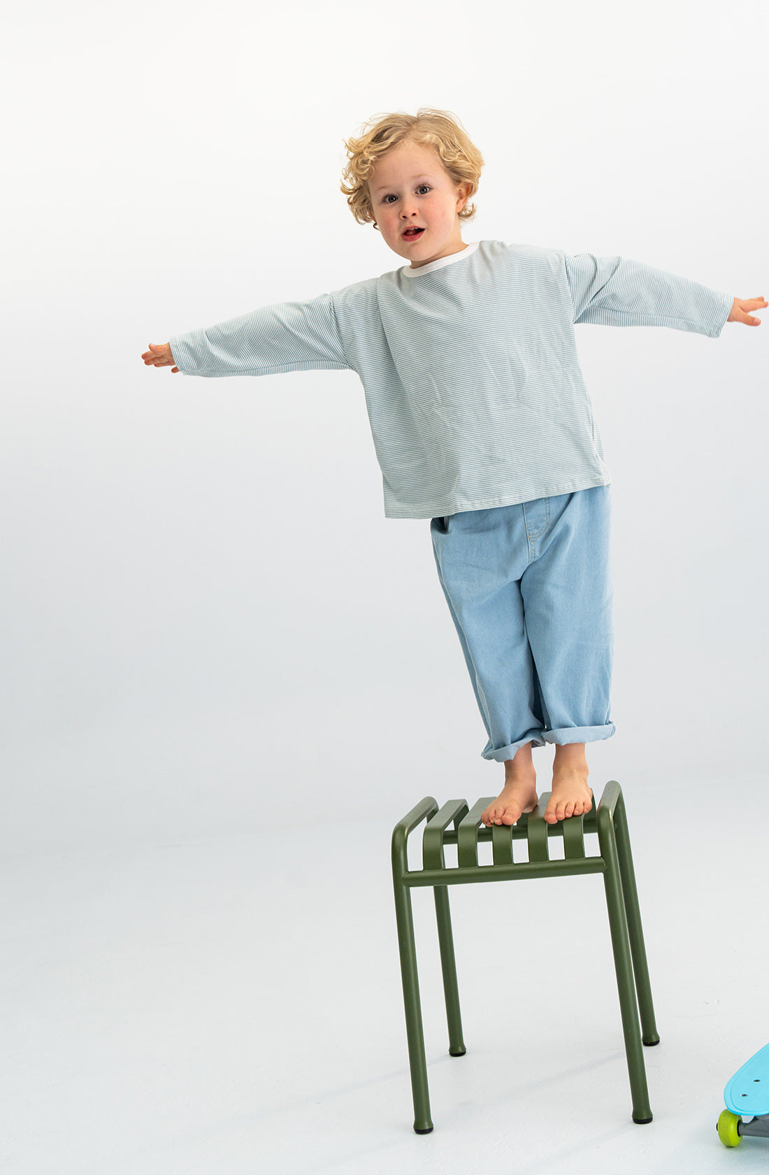 A young child with curly blonde hair stands barefoot on a green metal stool, wearing the SONNY LABEL Stripe Long Tee Blue and light blue pants. The child balances with arms spread out wide in a T-pose against a plain, light-colored background.