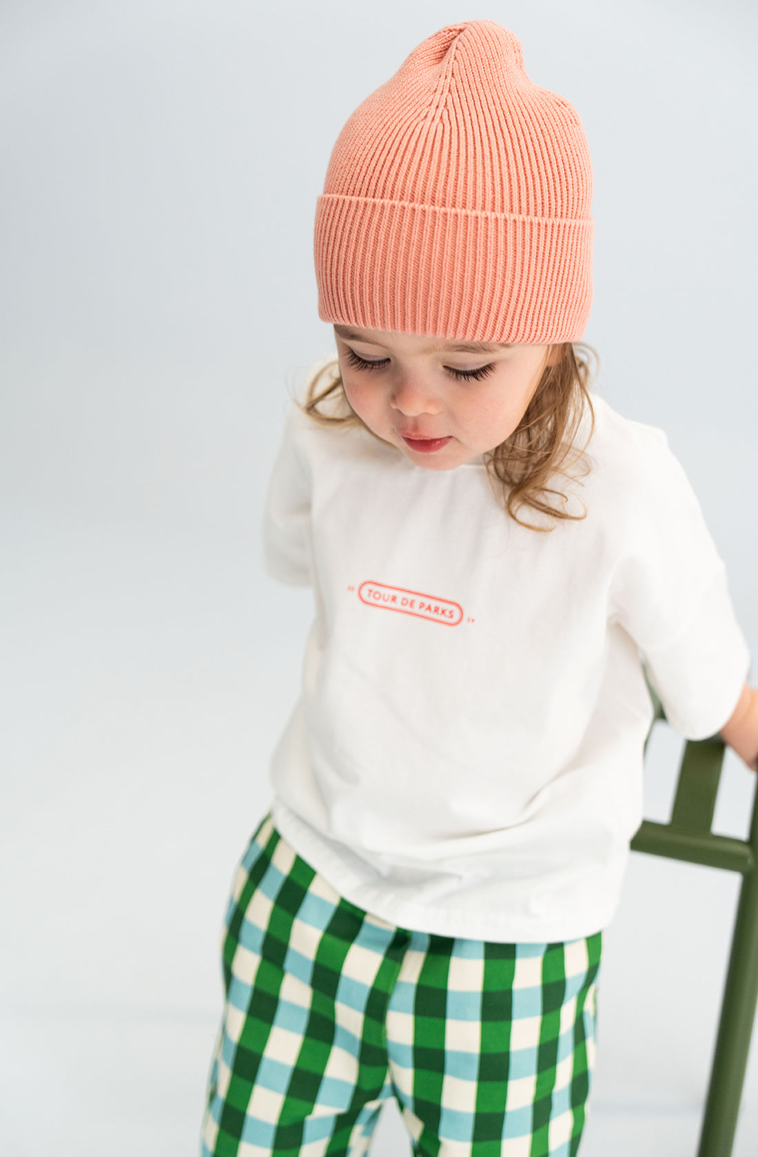 A toddler wearing a SONNY LABEL Cotton Beanie in pink, a white t-shirt with red text, and green and white checkered pants sits on a green chair against a light background. The child looks down with a slight smile.