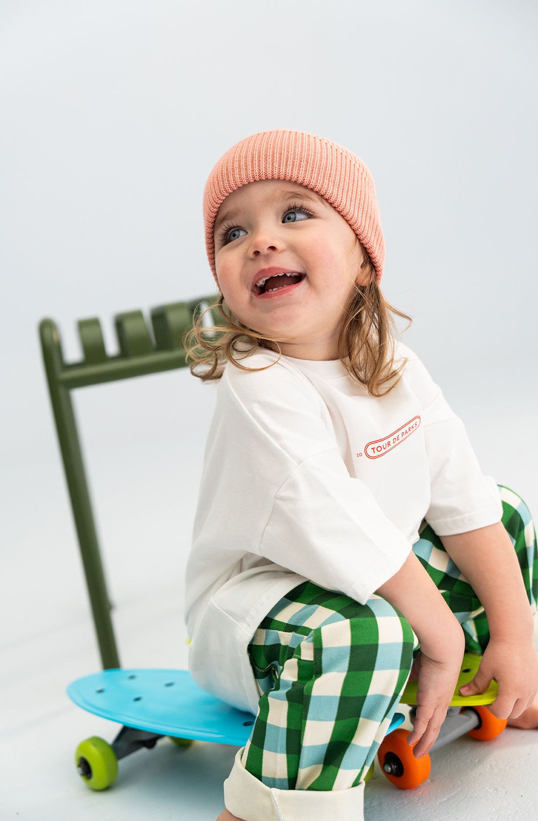 A young child with curly brown hair, wearing a chunky pink SONNY LABEL Cotton Beanie, a 100% cotton white T-shirt, and green-checkered pants, is smiling and looking up. They are sitting next to a colorful skateboard with a green metal structure in the background.