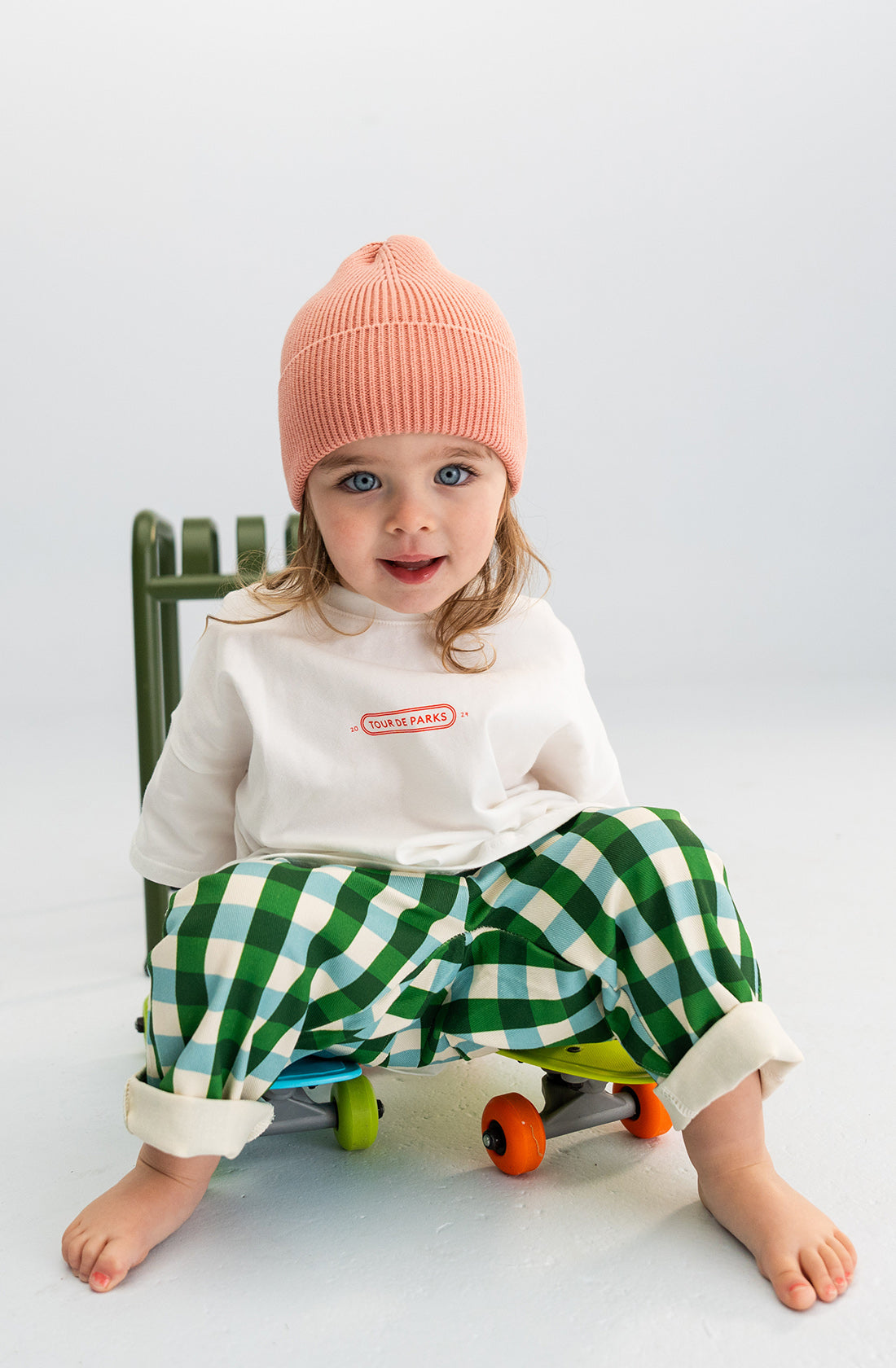 A toddler with long hair sits on a green chair, wearing a SONNY LABEL Cotton Beanie in Pink, a white long-sleeve shirt, and green-and-white checkered pants with the cuffs rolled up. The floor is white with a minimalist background as the toddler looks towards the camera.