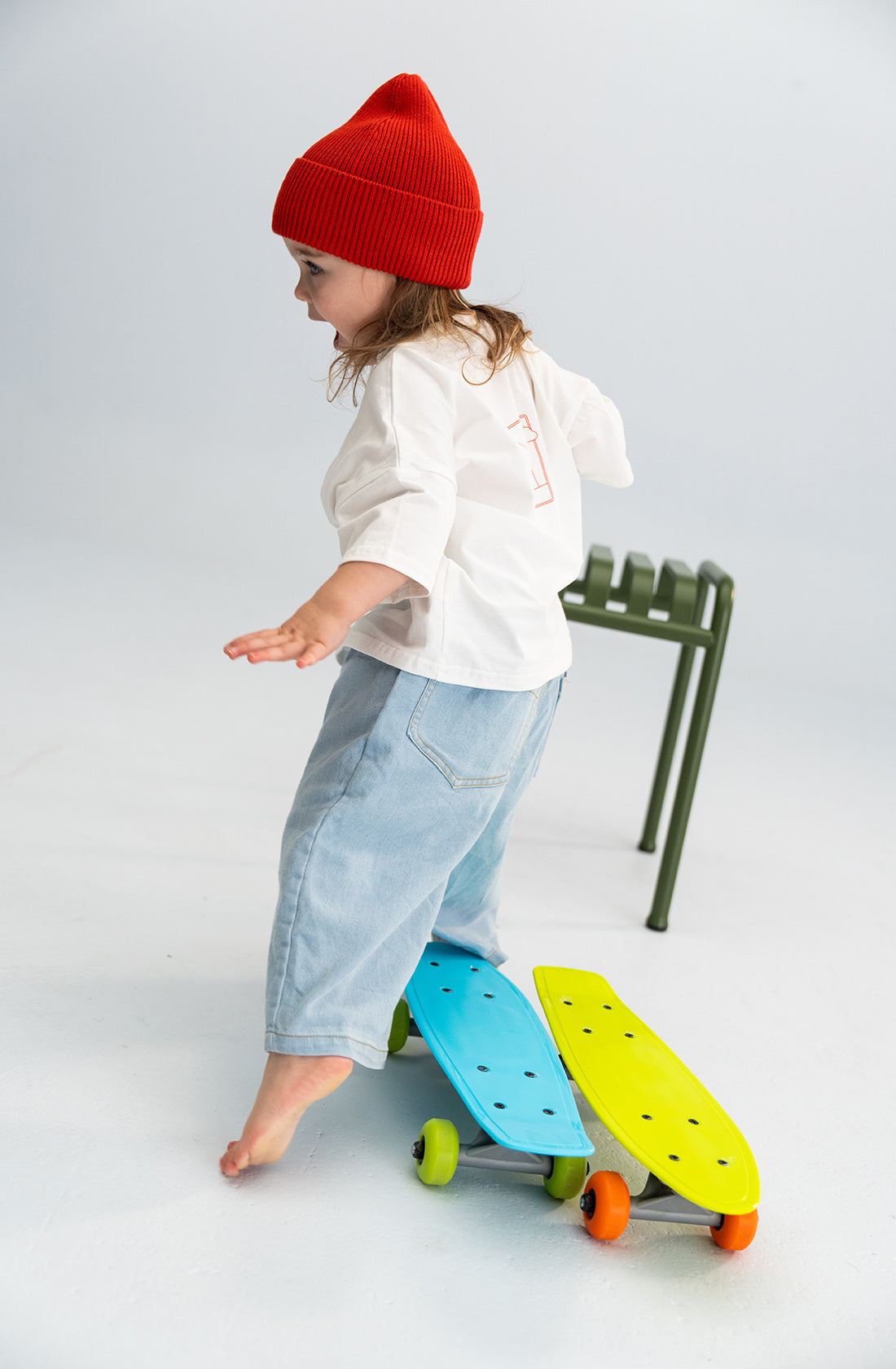 A young child with long hair wearing a red beanie, a white long-sleeve shirt, and Light Denim Jeans from SONNY LABEL is balancing with outstretched arms. The child is standing barefoot on a white surface with two colorful skateboards, one blue and one yellow, next to them.