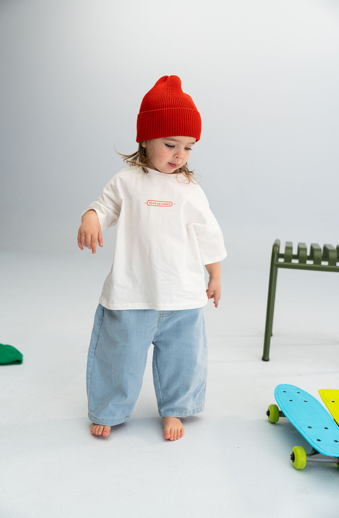 A small child wearing a red knit hat, a white t-shirt, and light denim jeans from SONNY LABEL with an elastic waistband stands barefoot indoors. Near them, a green chair and a colorful skateboard are visible on the floor. The background is a plain light-colored wall.