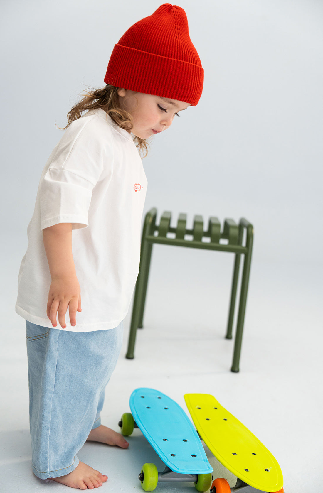 A young child with long hair, wearing a chunky red Cotton Beanie from SONNY LABEL, a white 100% cotton t-shirt, and light blue pants stands barefoot next to two small skateboards—one blue and one yellow. A green stool is in the background.