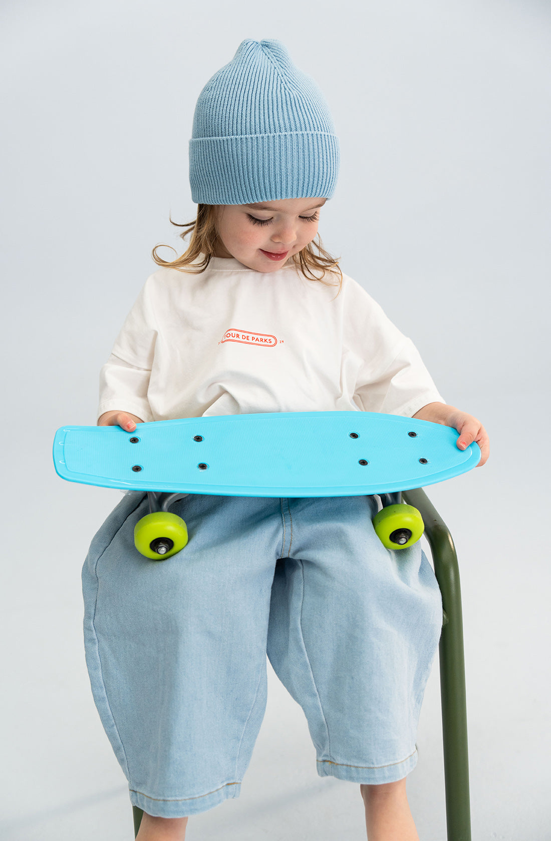 A young child sits on a green chair, holding a blue skateboard with bright yellow wheels. The child is wearing a chunky "Cotton Beanie Blue" by SONNY LABEL, along with a white shirt featuring red text, and light blue baggy pants. The background is set in a neutral, light-colored environment.