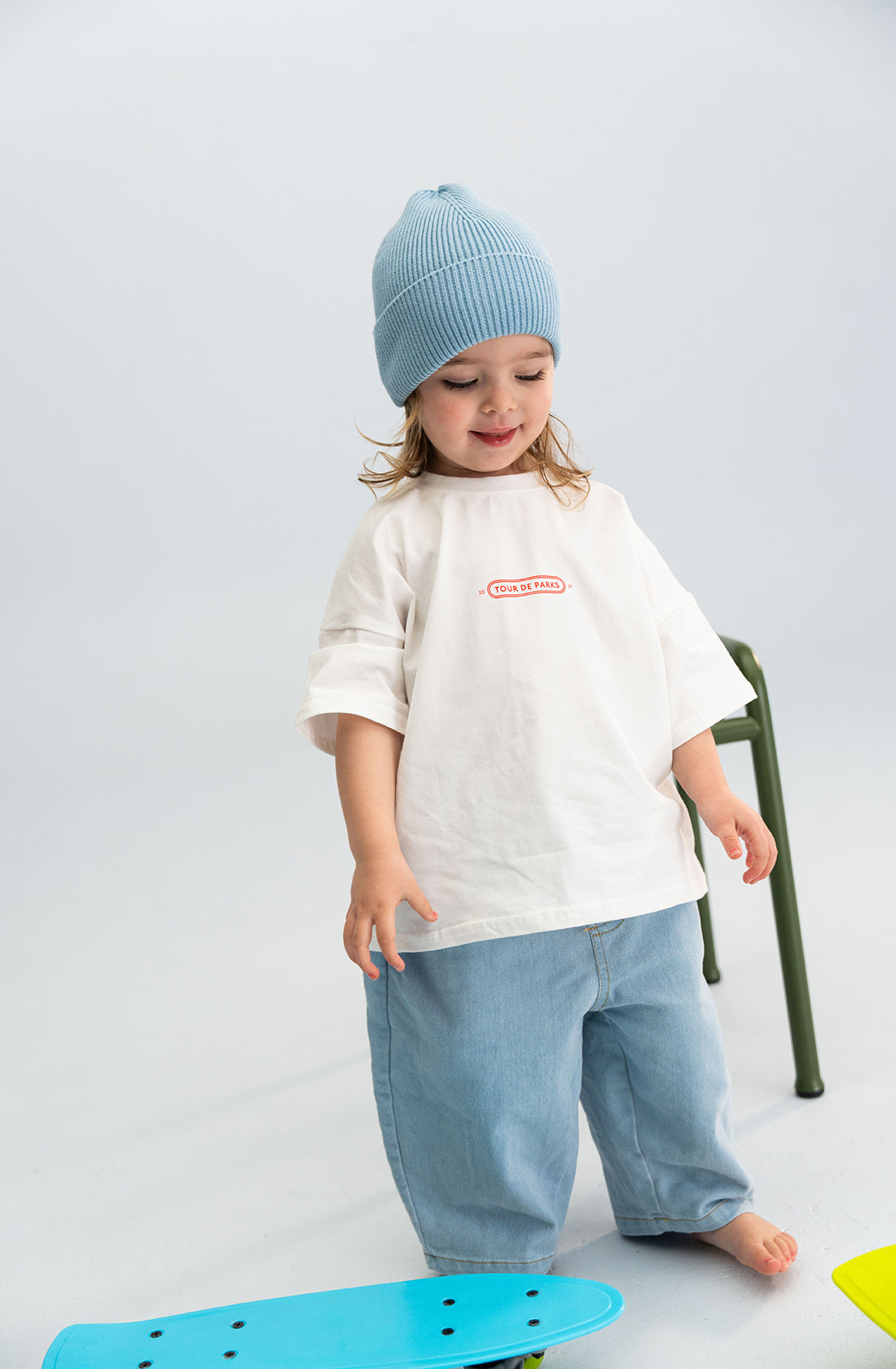 A young child wearing a SONNY LABEL Cotton Beanie in blue and an oversized white T-shirt with a red logo stands on a light-colored floor. They also wear loose-fitting blue pants. A colorful object, possibly a toy or skateboard, is on the floor near them.