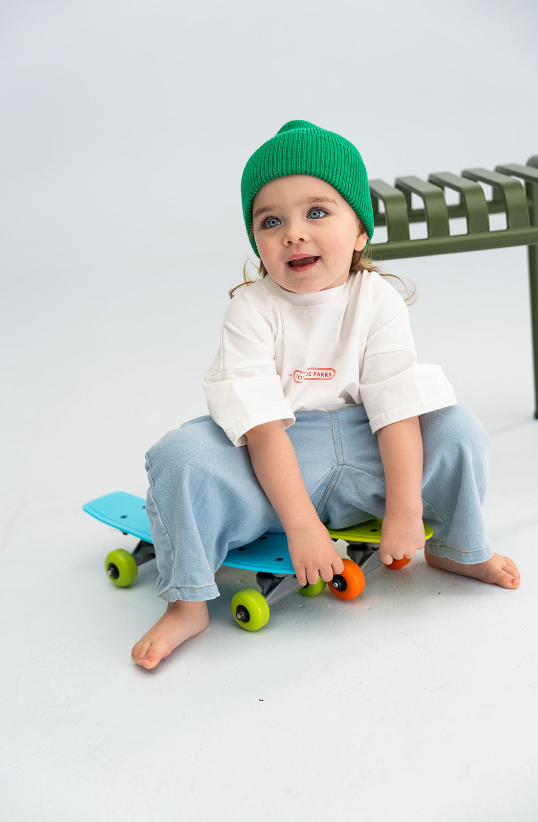 A young child with bright blue eyes sits barefoot on a blue skateboard, holding onto it with both hands. The child wears a white long-sleeve shirt, light blue jeans, and a chunky green Cotton Beanie from SONNY LABEL. They are seated on a white floor with a metal bench in the background.