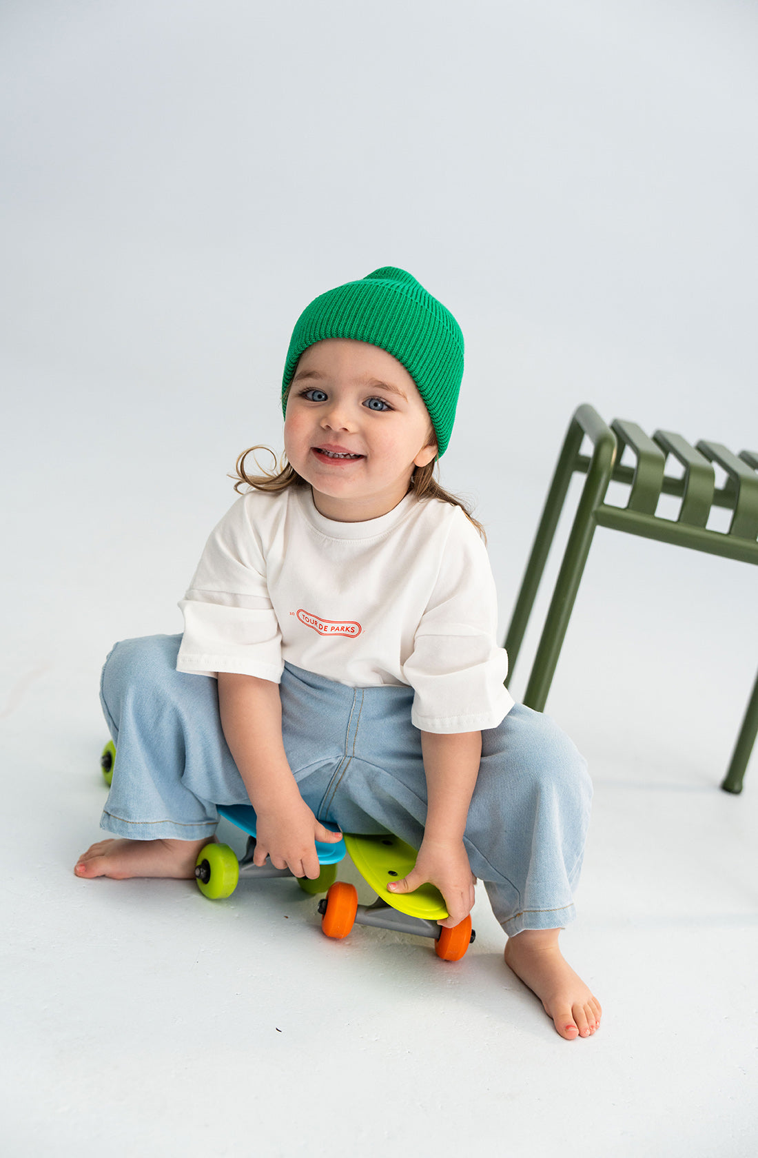 A young child wearing a chunky Cotton Beanie Green from SONNY LABEL, a white 100% cotton t-shirt, and blue jeans sits on a small, colorful skateboard indoors. They are smiling and appear to be having a good time. A green chair is partially visible in the background.