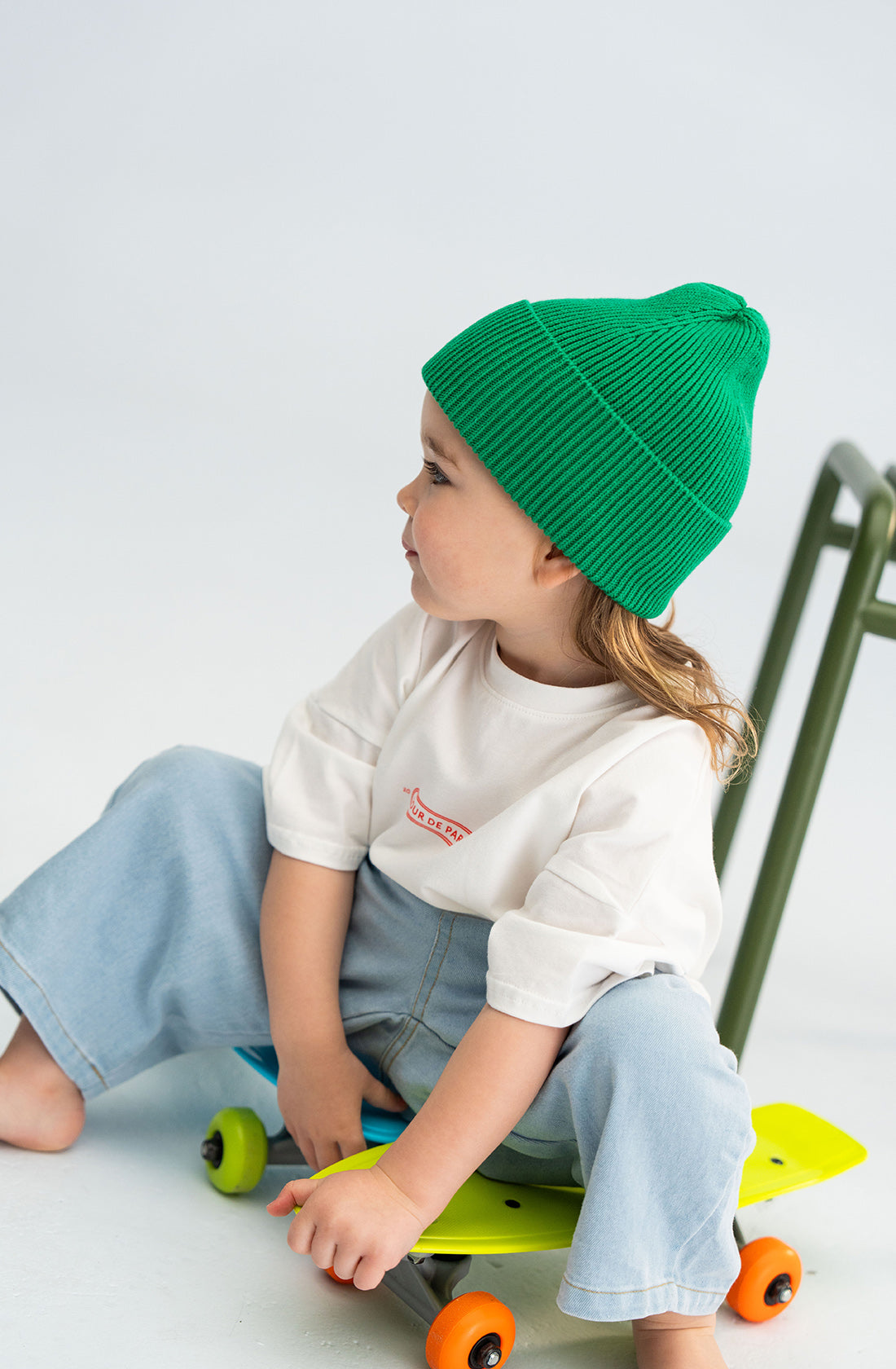 A young child with long hair wearing a bright green SONNY LABEL Cotton Beanie and a white shirt with rolled-up sleeves sits on a multicolored skateboard. The child is looking to the side, hands resting on their knees. A metal chair is visible in the background.