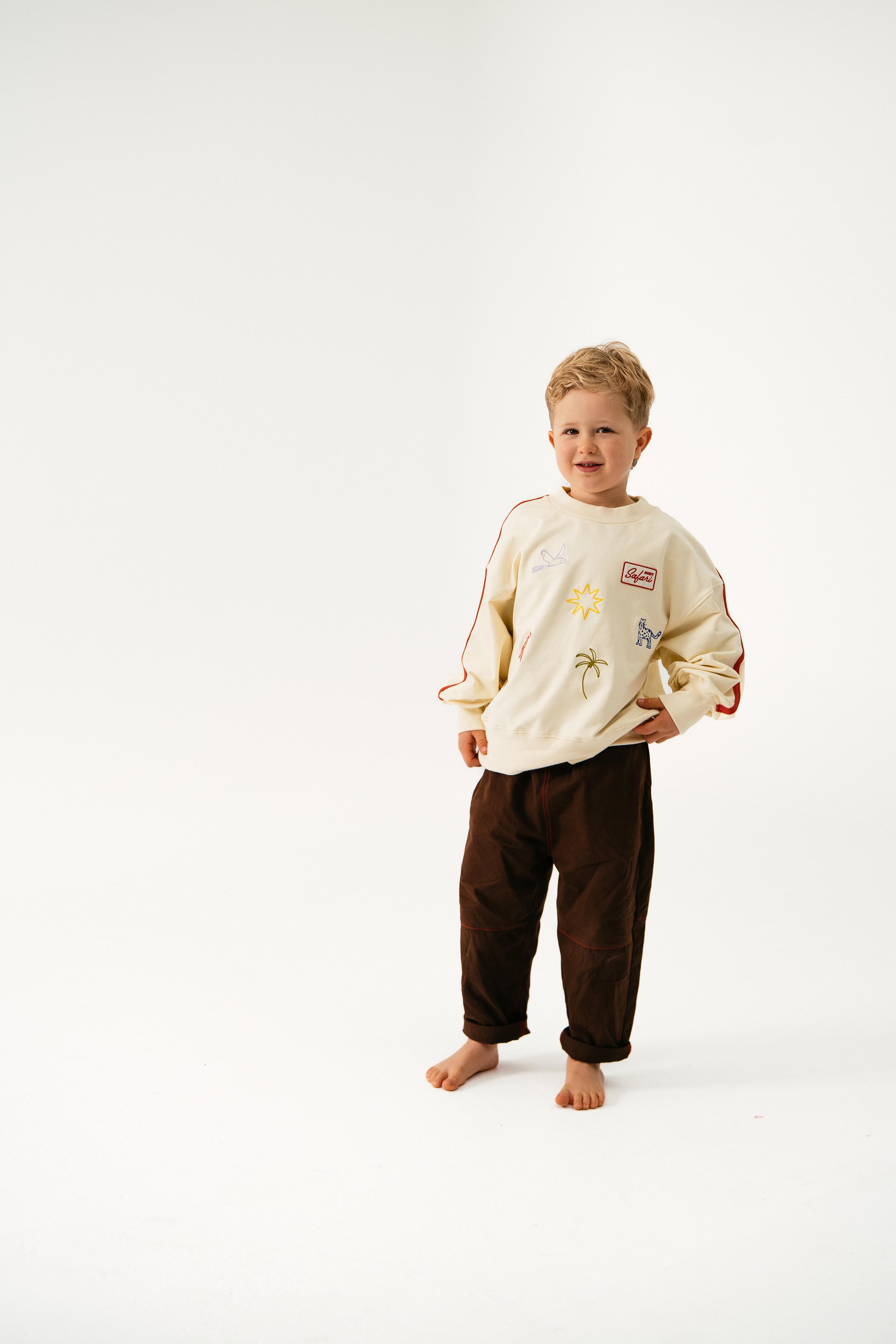 A young boy stands barefoot on a white background, smiling with one hand in his pocket. He wears a cream sweatshirt with colorful drawings and SONNY LABEL's brown Cargo Pants.