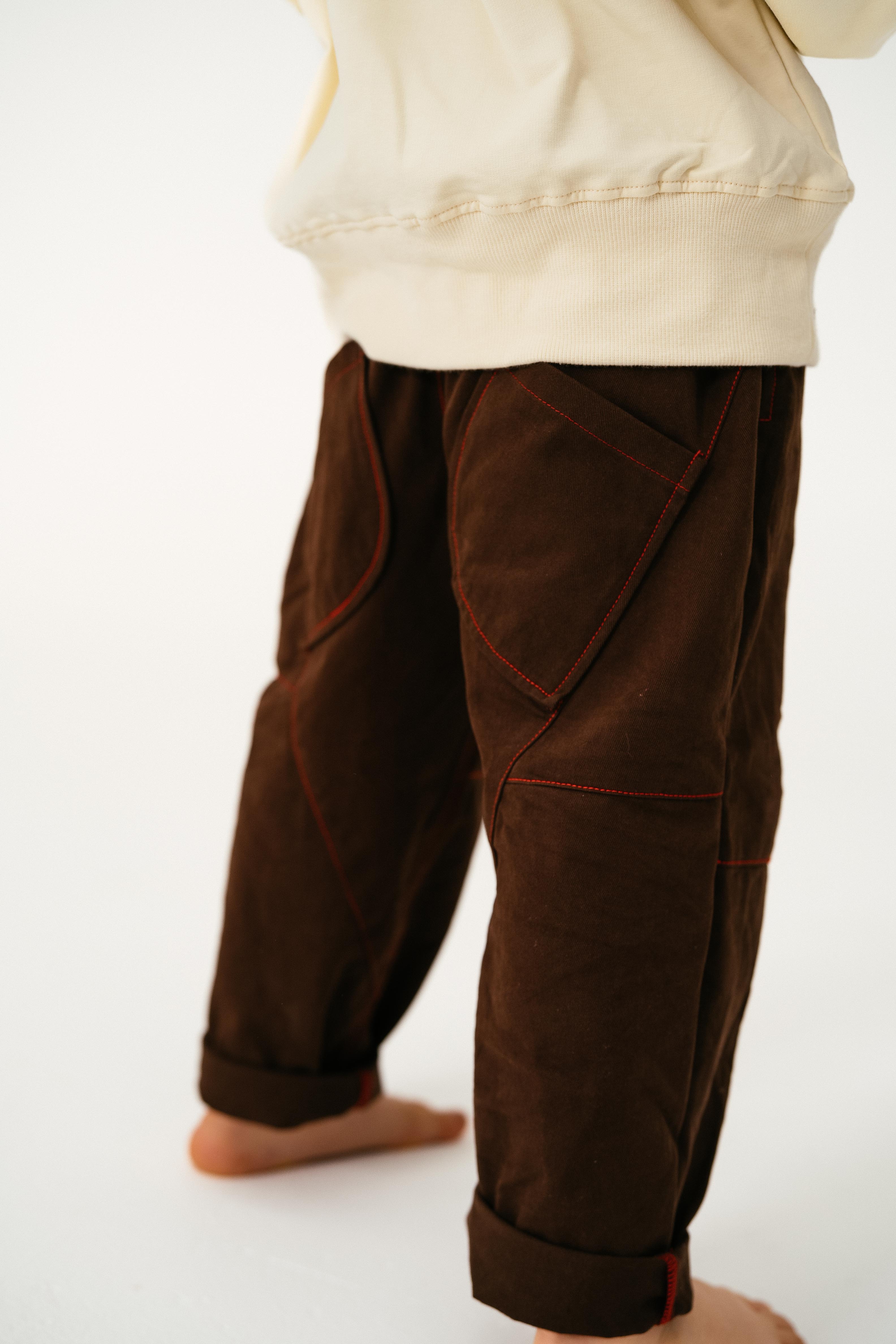 A child stands barefoot on a light surface, facing away, wearing SONNY LABEL’s dark brown Cargo Pants with red stitching and a cream sweatshirt.
