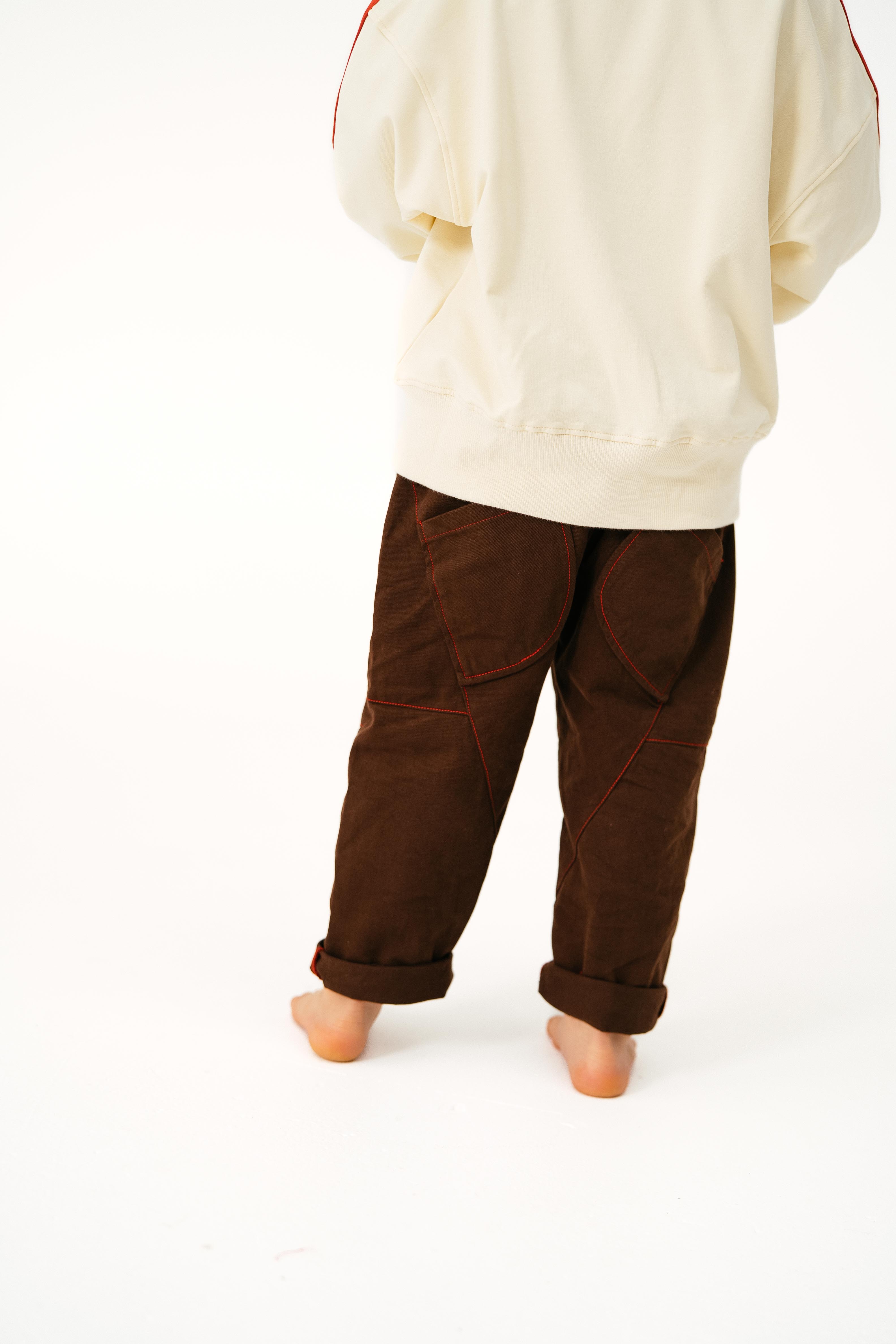A person stands barefoot on a white surface, wearing SONNY LABEL’s Sonny Label Cargo Pants in dark brown cotton twill with red stitching and loose legs, paired with a cream sweatshirt. The photo is taken from behind, showing only their lower body.