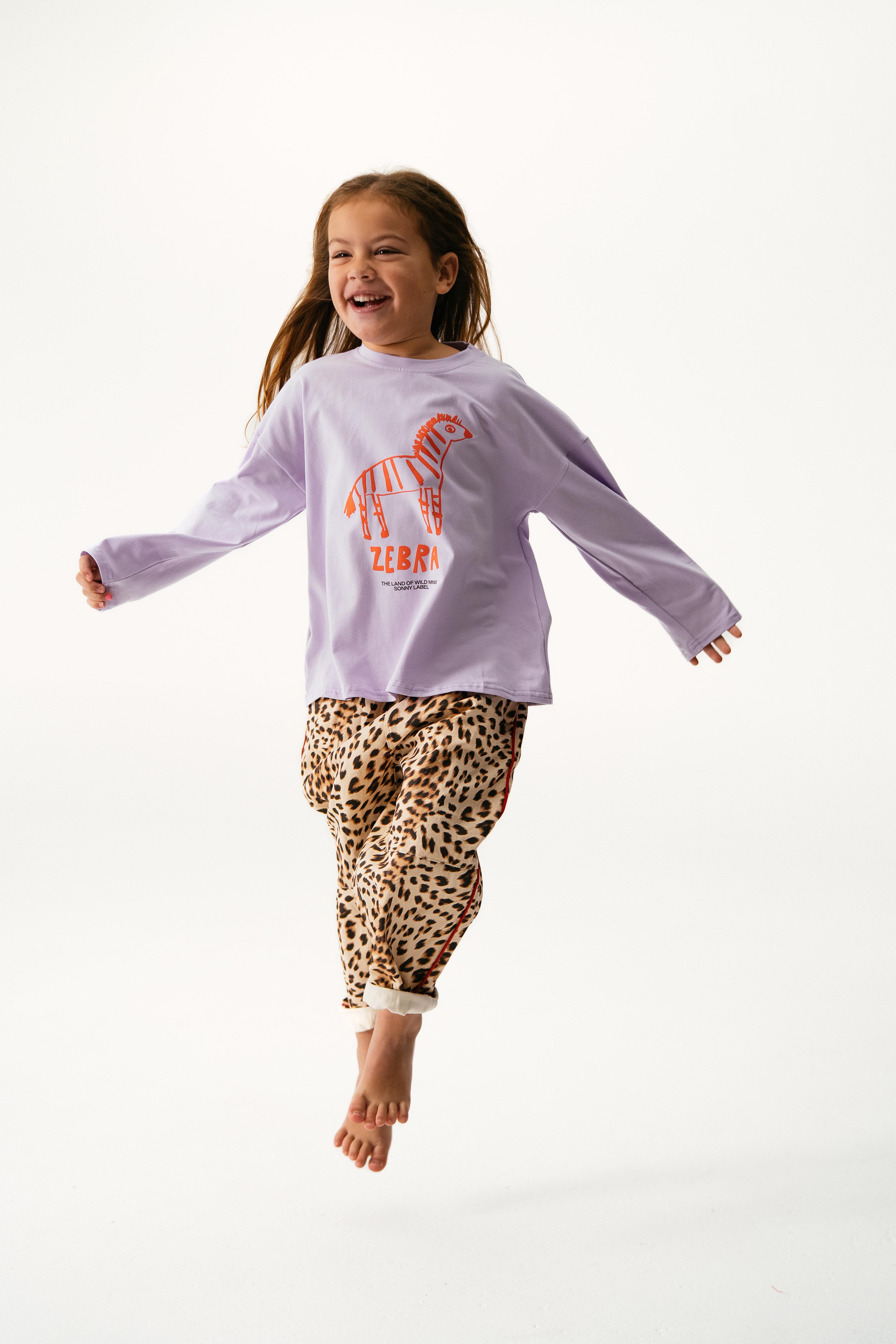 A smiling young girl jumps in the air wearing the SONNY LABEL Sonny Label - Animal Long Tee Lilac, featuring a zebra graphic and "ZEBRA" text, paired with leopard print pants against a plain white background.