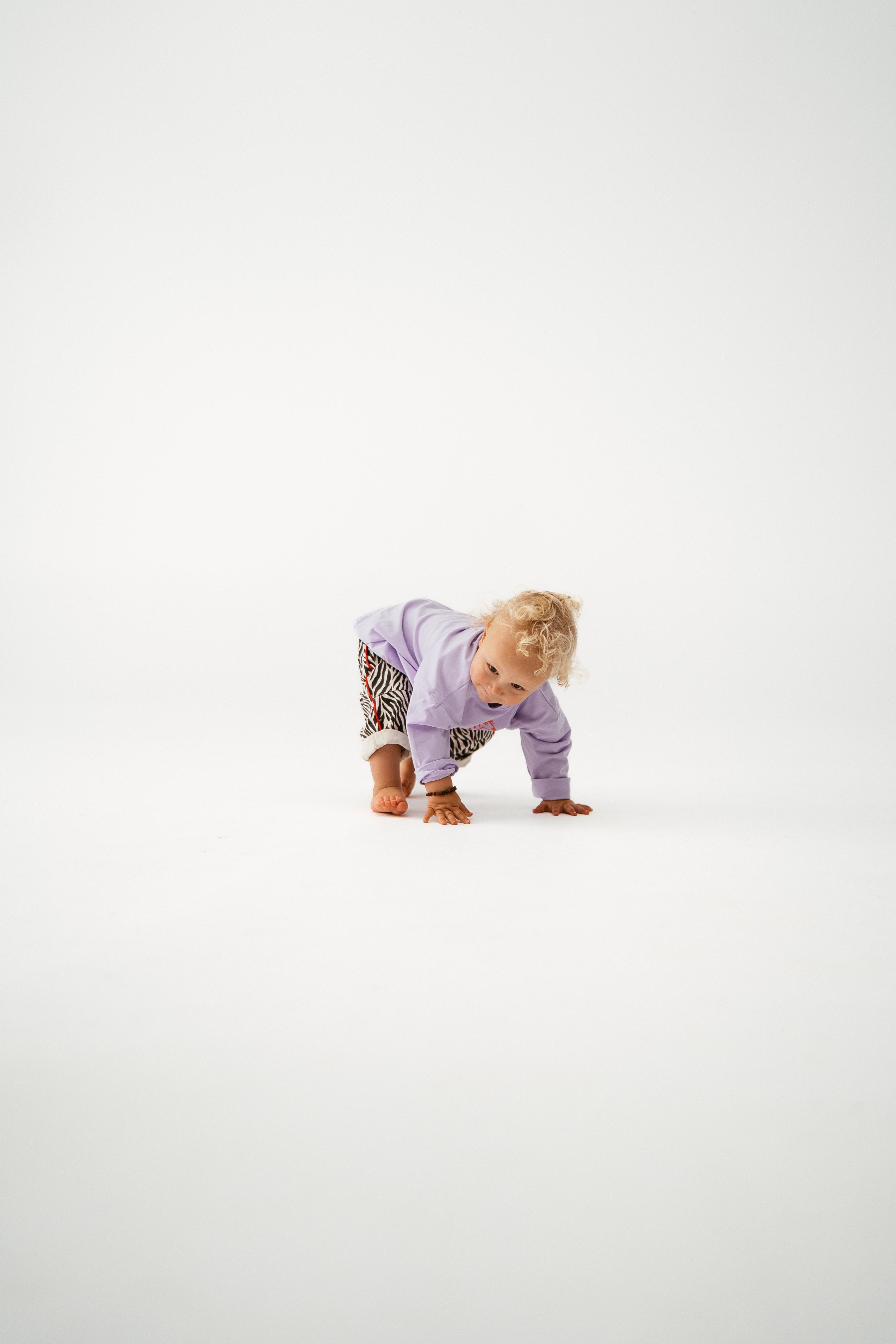 A young child with curly blond hair wears a light purple top and SONNY LABEL Sonny Label - Animal Twill Pants Zebra, bending down to touch the floor against a plain white background.