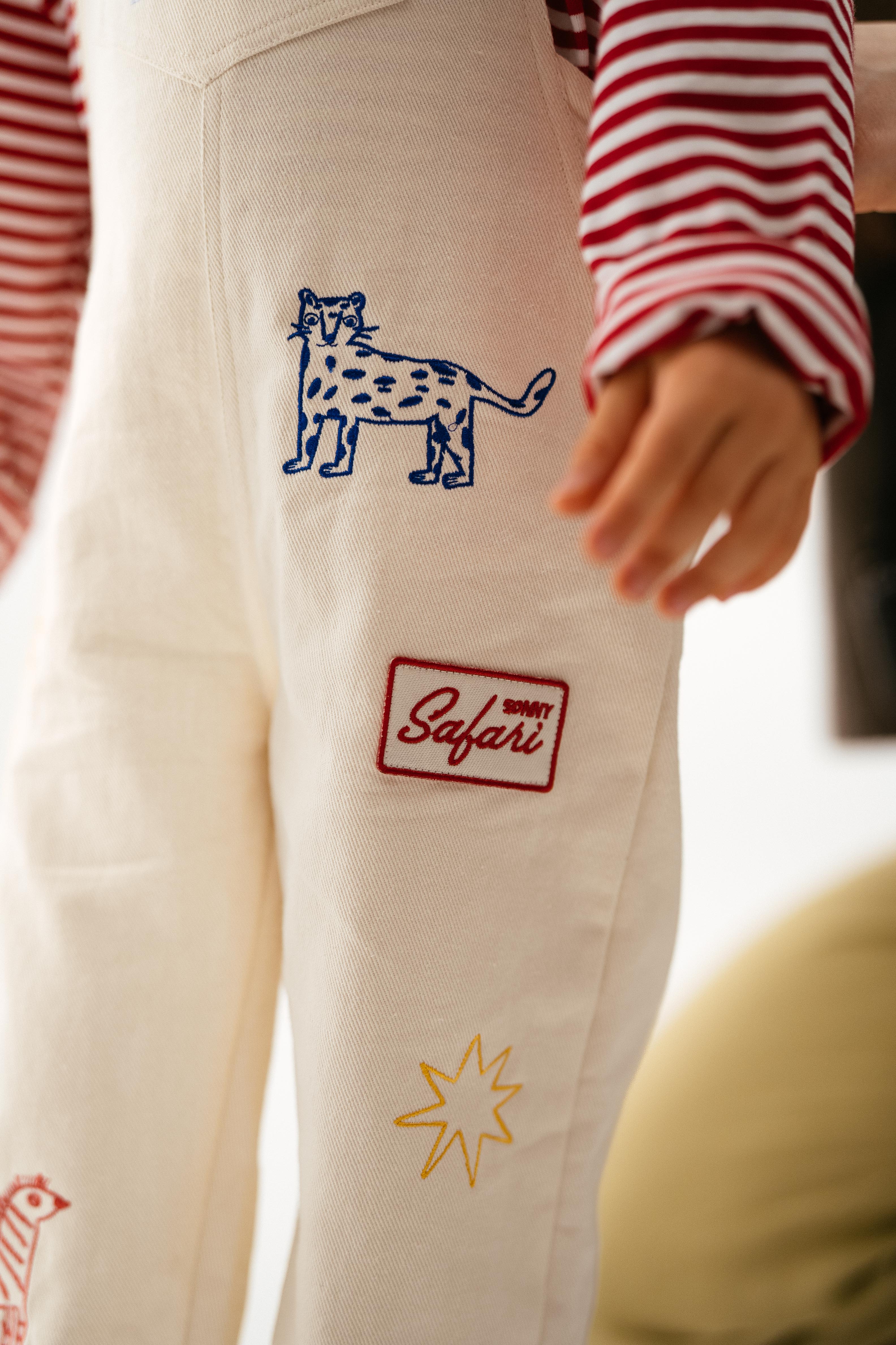 A child wears SONNY LABEL’s Sonny Label Dungarees—cotton twill, cream-colored, with embroidered animals and a bold Safari patch—paired with a red-and-white striped long-sleeve shirt.