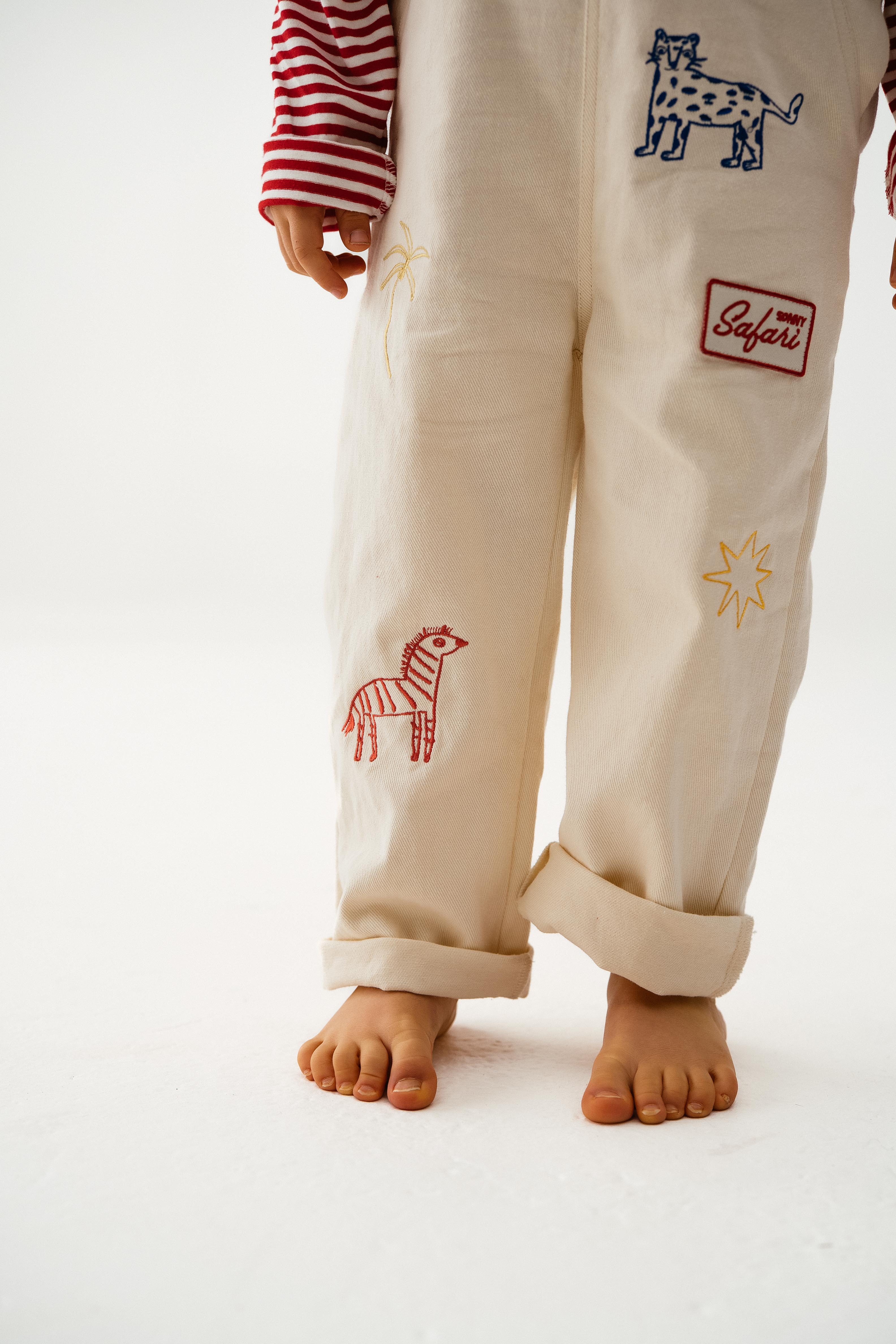A child, head not visible, stands barefoot on a white surface wearing SONNY LABEL's Sonny Label Dungarees with embroidered animals and safari patches over a red-and-white striped long-sleeve shirt.