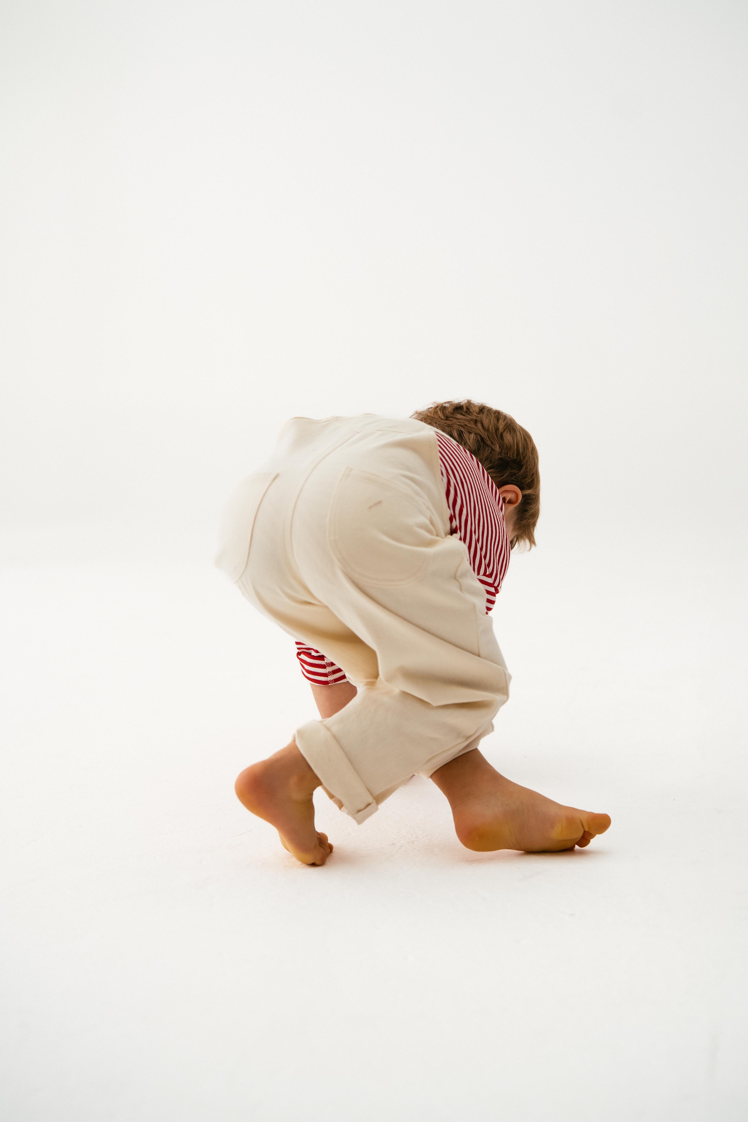 A young child wearing SONNY LABEL’s "Sonny Label - Dungarees" cream cotton twill pants and a red-and-white striped shirt bends over barefoot on a white background.
