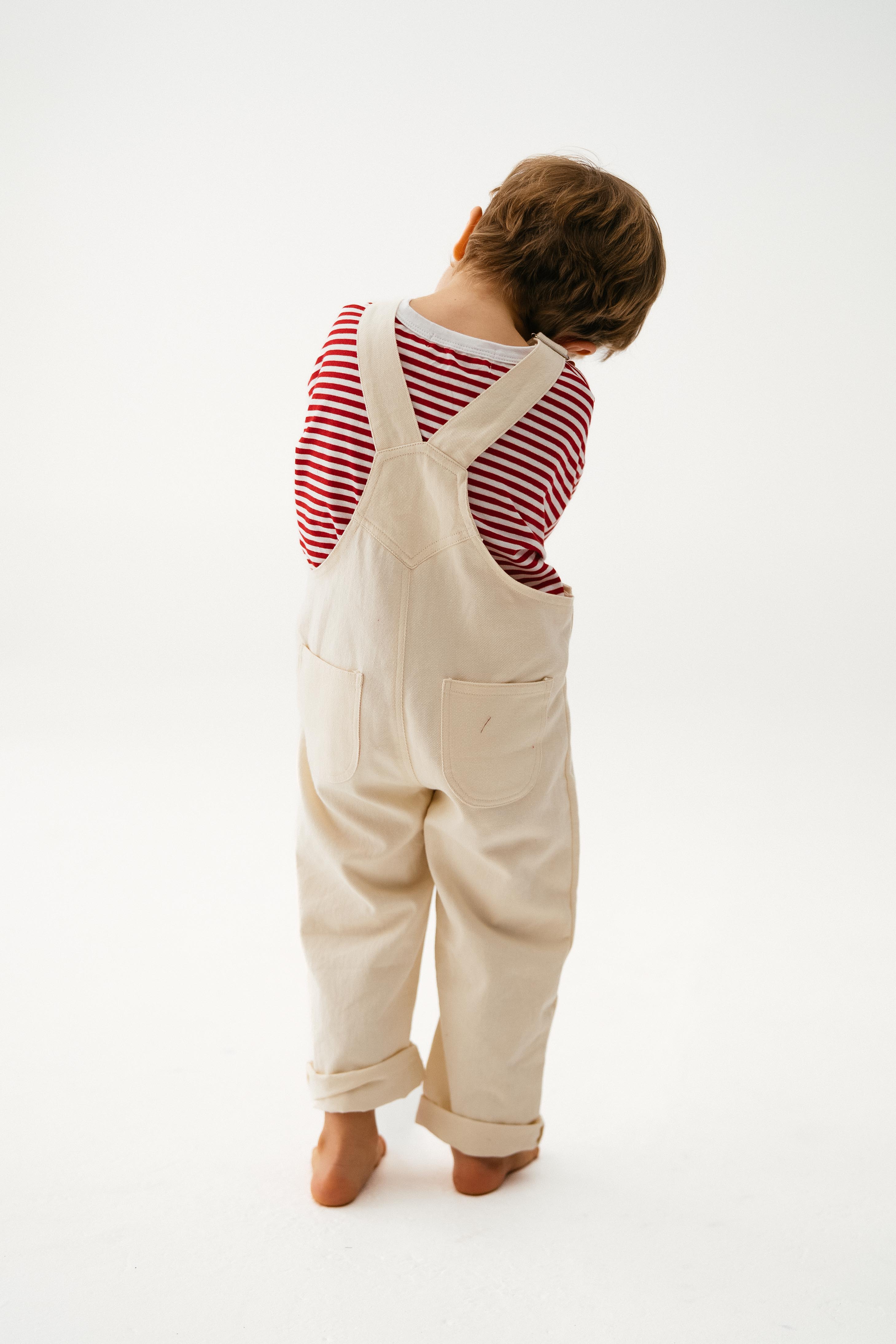 A young child stands barefoot on a white backdrop, facing away, wearing SONNY LABEL's Sonny Label Dungarees over a red and white striped long-sleeve shirt, head tilted to the side.