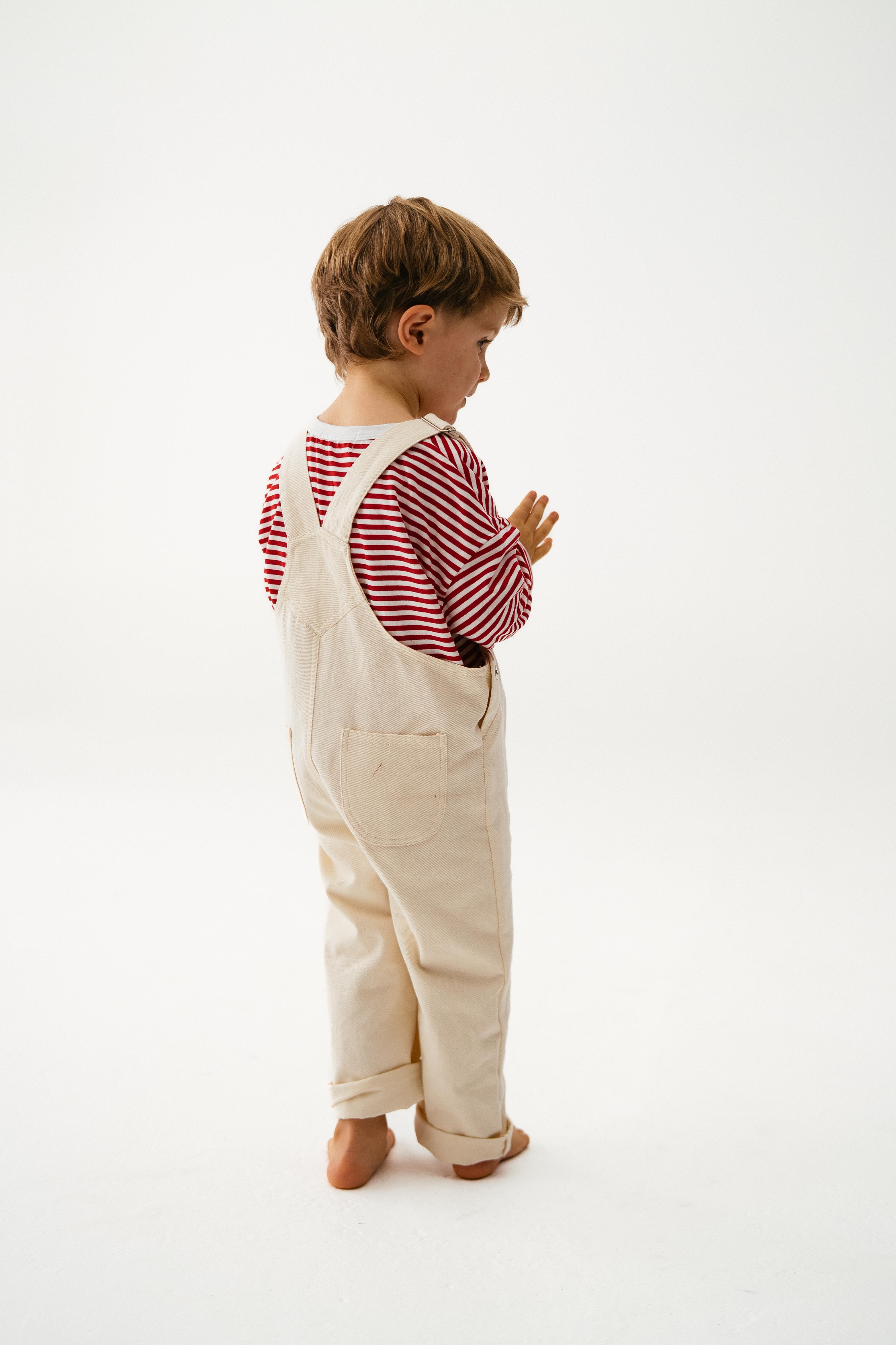 A young child in SONNY LABEL’s Sonny Label Dungarees and a red-and-white striped long-sleeve stands barefoot, slightly turned away, on a plain white background. The light cotton twill overalls feature a playful safari patch.